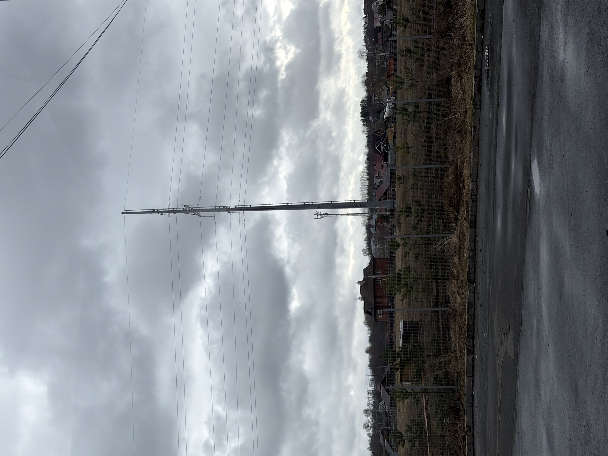 The image shows a street in Izhevsk, Russia. Overhead power lines are visible, supported by a tall metal tower. Adjacent to the road is a wire fence that separates the road from the properties beyond. These properties contain a mix of houses and vegetation. The road itself appears wet, possibly from recent rain. The sky is overcast with a cloudy appearance.