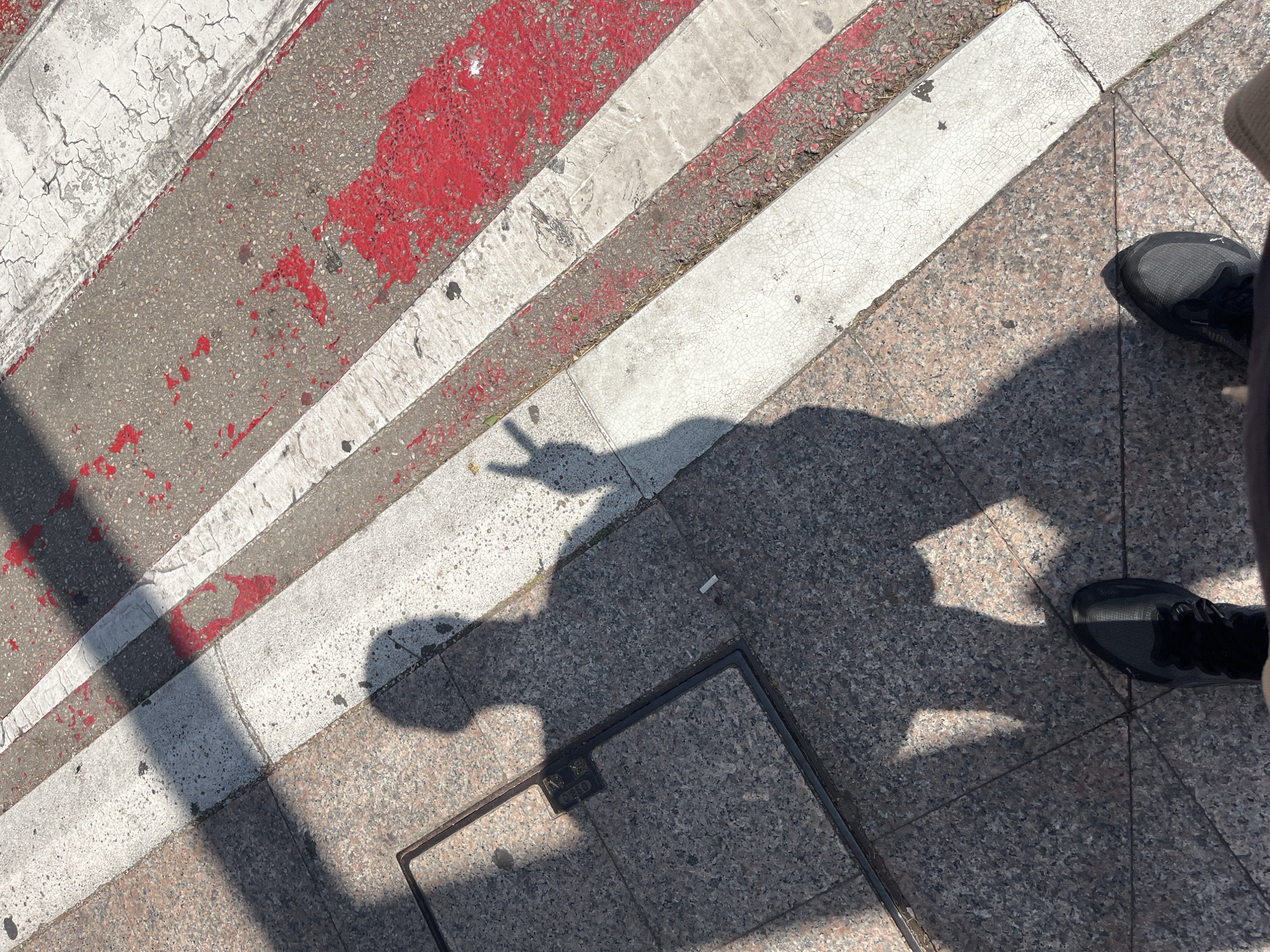 The image shows a pedestrian crossing on a street in Budva, Montenegro. The crossing has white stripes painted across the asphalt, with red paint markings also visible on the road surface. In the foreground, two feet wearing black athletic shoes are visible, suggesting the perspective of someone standing on the sidewalk. A shadow of a person is cast on the ground, with the shadow's hand forming a peace sign. To the left of the shadow, a square metal manhole cover is embedded in the granite-tiled sidewalk. The scene is illuminated by sunlight, creating distinct shadows.