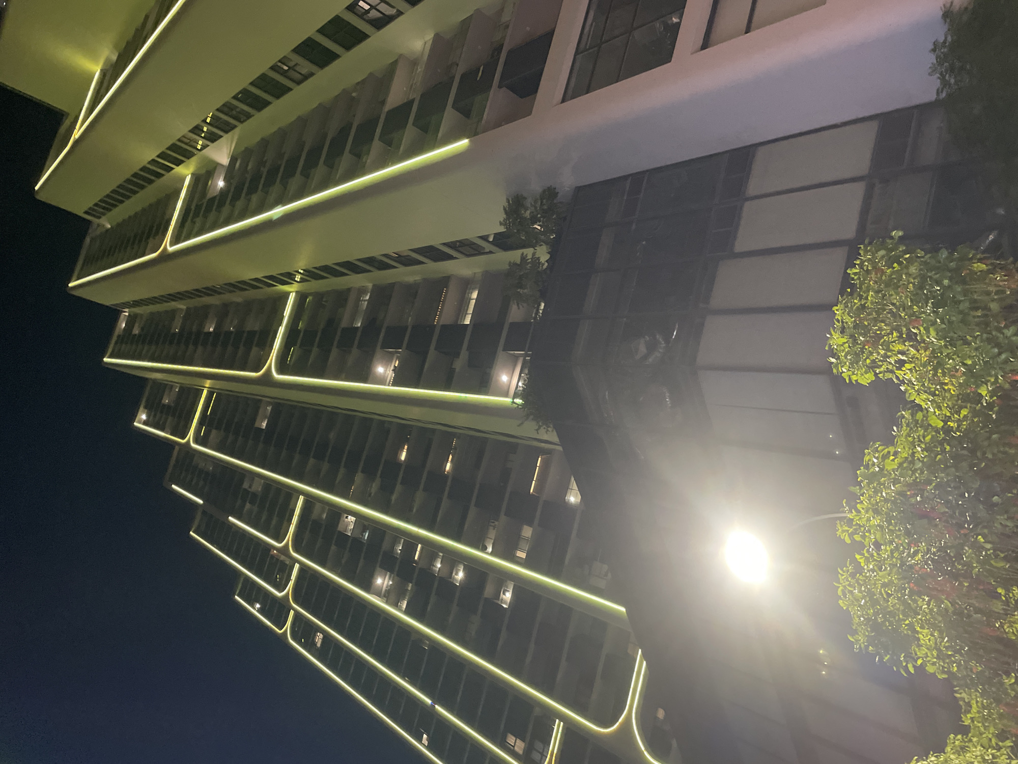 The image displays a modern high-rise building at night, dramatically illuminated by linear yellow-green LED lights that highlight its architectural design. The building features multiple levels with pronounced, possibly cantilevered, floor plates, each outlined by these bright light strips. Its facades are composed of dark glass and lighter-colored panels, reflecting some of the ambient light.
To the right side of the frame, several trees with dense green foliage are visible, some partially lit by the building's glow. A very bright streetlamp dominates the lower right foreground, emitting a powerful white light that creates a prominent lens flare and illuminates parts of the surrounding foliage. The sky is dark, indicating nighttime, and the scene is devoid of people or discernible activity, lending a calm and contemporary atmosphere.
The architectural lighting gives the building a distinctive and futuristic appearance against the dark backdrop. This scene is located in Bắc Nha Trang, Vietnam. No visible text is present in the image.