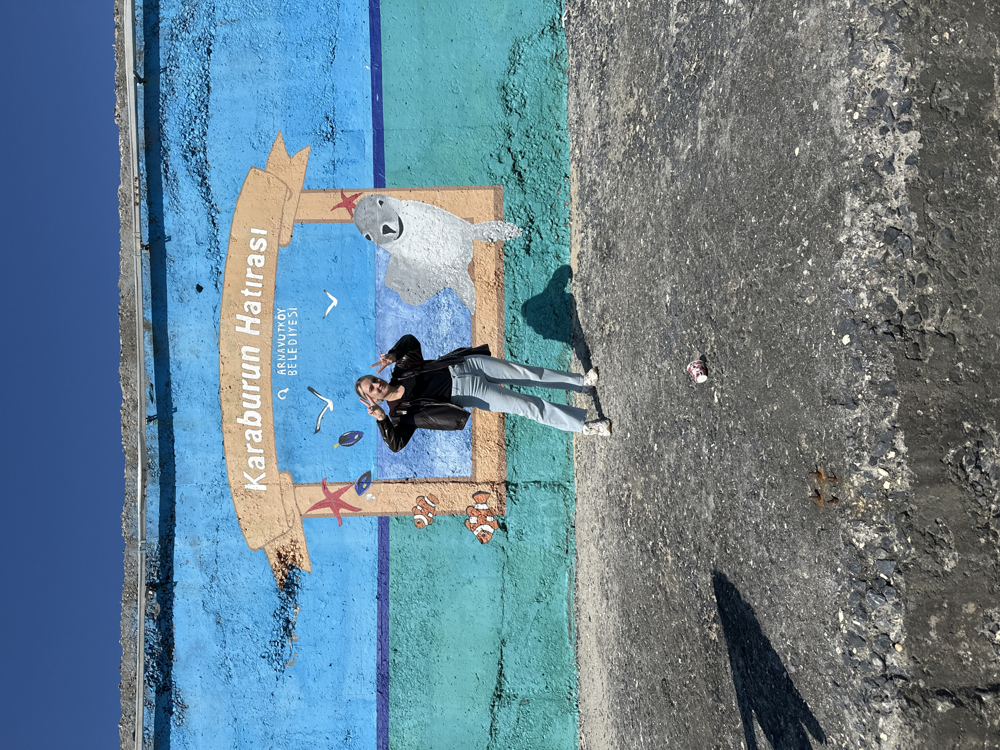 A young woman in a black jacket and light blue jeans poses with a peace sign in front of a painted mural in what can be inferred as Arnavutköy, Turkey. The mural is painted on a concrete wall, with the top portion of the wall painted blue to mimic the sky and the lower portion painted various shades of blue and green to mimic water. The mural features a painted banner with the words "Karaburun Hatırası" and "ARNAVUTKÖY BELEDİYESİ" on it. In the painted water, there is a seal, two clownfish, a starfish, and a seagull. The ground adjacent to the wall is asphalt and there is a used coffee cup on the ground. The sky is visible above the wall, indicating the photo was taken outdoors during daylight hours.
