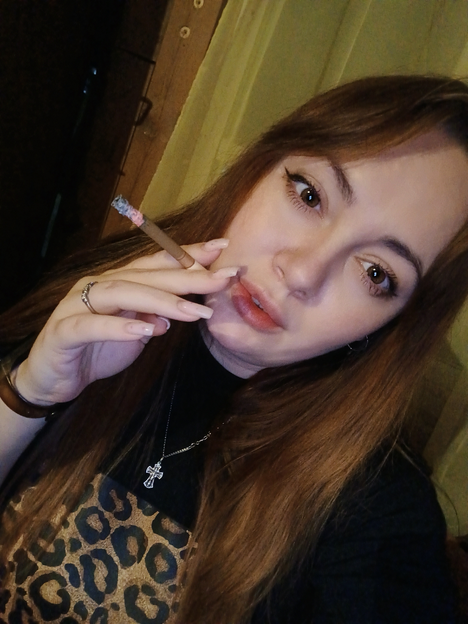 The scene depicts a female individual in a dimly lit indoor setting. She has long, reddish-brown hair and brown eyes with visible eyeliner. Her lips are slightly parted and tinted red. She is holding a burning cigarette between the index and middle fingers of her left hand; the cigarette's ash tip shows pink and blue hues. A silver-colored ring with small stones is on her left ring finger, and her fingernails are long and appear to have a clear or nude polish. A dark-colored bracelet is present on her left wrist. She is wearing a dark top, beneath which or as part of which, a garment with a leopard print pattern is visible. A silver-colored necklace with a crystalline crucifix pendant rests on her chest. The background features a vertical wooden panel or beam to the upper right, adjacent to a light-colored, possibly yellowish-green, curtain or fabric. The individual is looking directly at the viewer. The scene is located in the city of Ольховка, Russia.