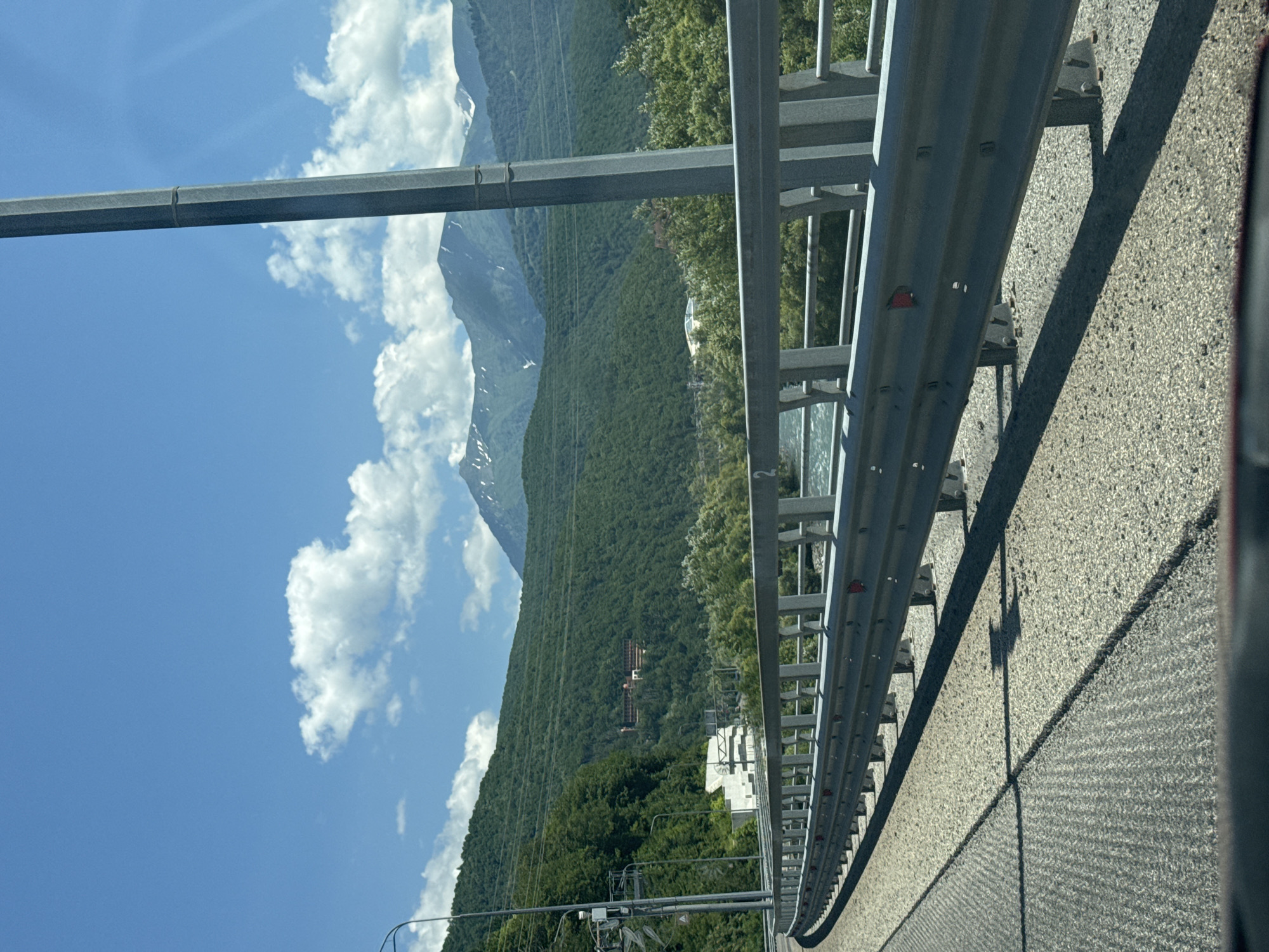 A view from a car traveling on a road in Krasnaya Polyana, Russia, shows a mountainous landscape under a clear blue sky with scattered white clouds. The road curves to the left, bordered by a metal guardrail. In the distance, lush green forested mountains rise, with some patches of snow visible on the peaks. A river flows below the road, and a large, multi-story building complex is nestled into the side of one of the mountains. A tall, grey pole, likely a street light or utility pole, stands prominently in the foreground on the right side of the frame. The road surface is paved with asphalt and gravel.