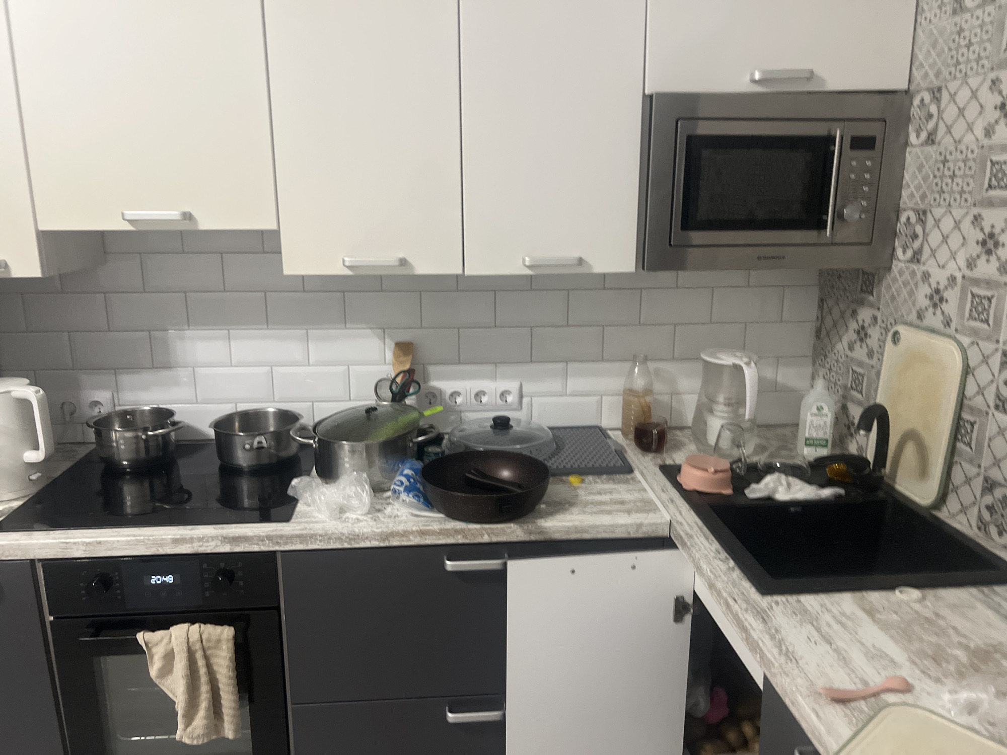 The image depicts a cluttered kitchen counter. On the counter, there are various kitchen appliances and cookware, including pots, pans, a kettle, and a microwave. The cabinets above the counter are white and modern. Below the counter, there is a dark-colored oven with a towel hanging from it. The overall impression is one of a well-used and somewhat disorganized kitchen space. The specific location, "Veteran-2, Russia," is mentioned, but there are no visual cues in the image to support or contradict this location. The image is a still photograph, so no actions or interactions are depicted beyond the static arrangement of objects.