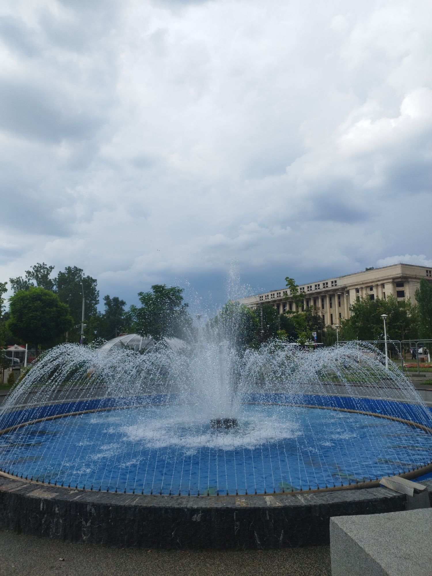 A fountain is shown in the foreground, with water spraying in a dome shape. The fountain has a blue basin and a dark stone base. Behind the fountain, there are trees and a large, light-colored building with columns. The sky is cloudy and gray. The scene is located in Bucharest, Romania, specifically near the Romanian Athenaeum.