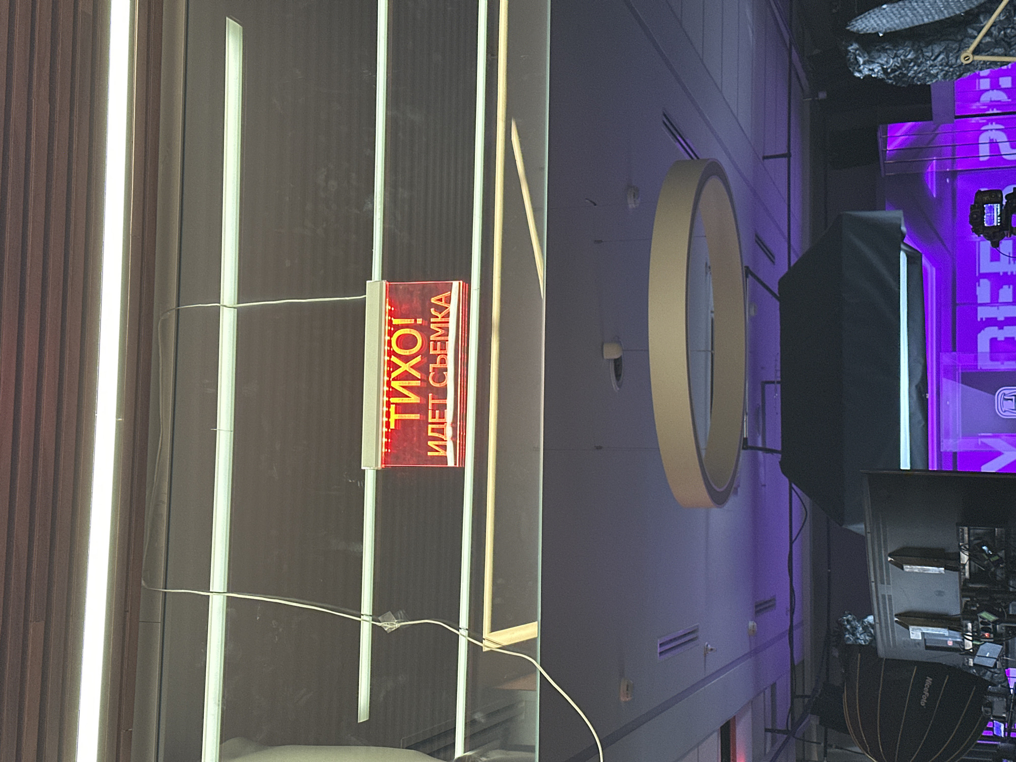 A vertical eye-level shot shows an indoor scene, including signage in a language other than English, set up lights, and wall decor. In the center of the frame, a rectangular sign with a red background displays writing that appears to read "ТИХО! ИДЁТ СЪЁМКА" in a white, vertical font. The sign is attached to a glossy surface with vertical lines and parallel, long, thin illuminated light fixtures.
To the right, a large circular ornament is visible on the wall. The walls are illuminated with a bluish-purple lighting, and there is photography equipment, like lights and reflectors, set up. In the background, bright purple lighting is visible and possibly a screen or display with lettering. Overall, the image indicates a photoshoot is in progress. The exact location in the city of Unknown, Unknown is not discernible from this image.