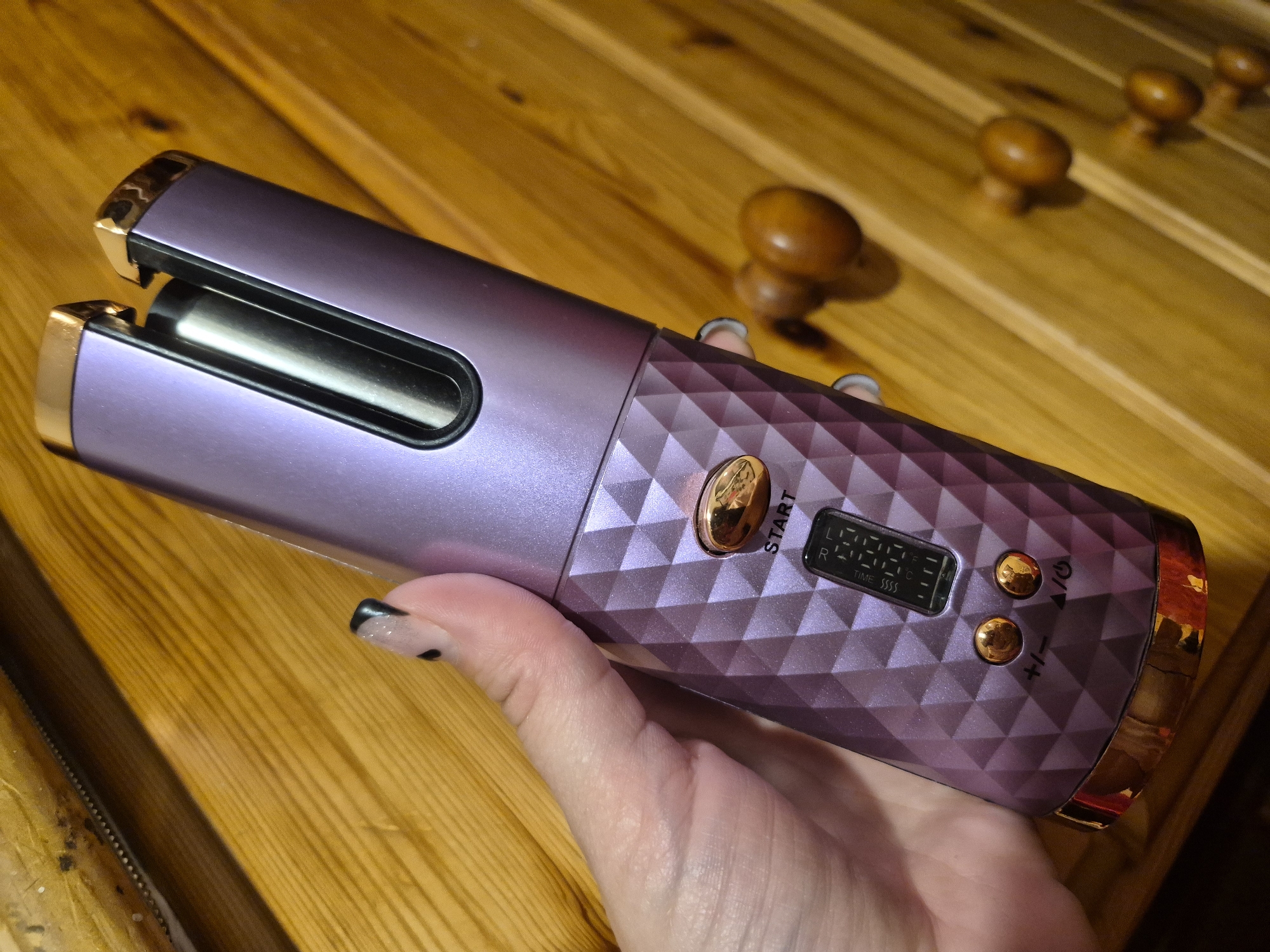 A close-up shot shows a person holding a purple automatic hair curler with gold accents against a backdrop of a wooden surface. The hair curler is cylindrical with a diamond-patterned body and a gold-colored opening at the top. It features a small digital display and several gold buttons, including a "START" button and buttons labeled with "+/-" and a power symbol. The person's hand, with a black and white speckled fingernail, is visible holding the device. The wooden surface appears to be part of a piece of furniture, possibly a desk or dresser, with visible grain and texture. The lighting suggests an indoor setting, and the overall image quality is clear, indicating a photograph taken with a modern camera. There is no discernible information within the image to indicate a specific location in Sandwell, United Kingdom, or any urban features.