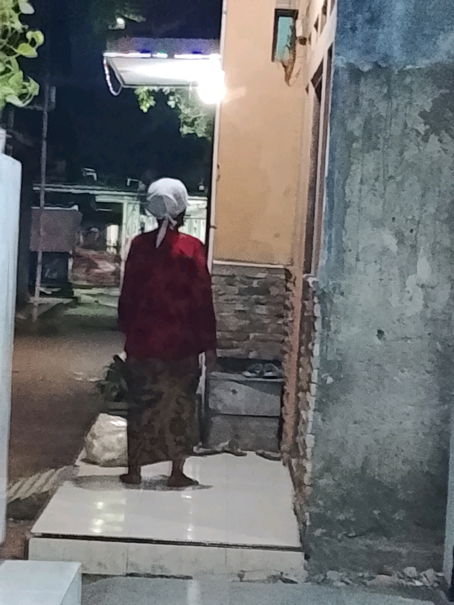The image shows a person from behind, wearing a red long-sleeved shirt and a patterned skirt. They have a white headscarf tied at the back. The person is standing on a white tiled platform in front of a building with a textured, light brown wall and a blue-grey wall to the right. There is some greenery on the left side of the image and a bag or bundle near the person's feet. The scene appears to be outdoors in a somewhat unlit environment, possibly at dusk or night, with artificial lights illuminating the area.