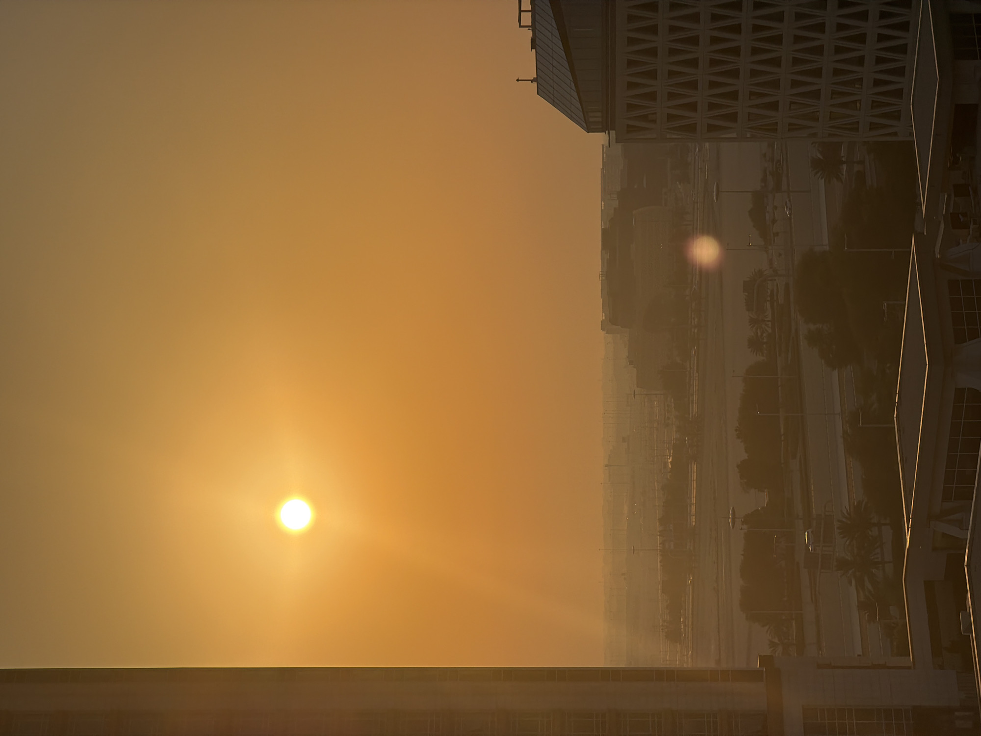 The image shows a city skyline at sunrise or sunset. The sun, a bright white orb, is positioned on the left side of the frame, casting a warm, golden glow across the hazy sky. The sky transitions from a bright yellow near the sun to a softer orange and then to a muted brown towards the horizon.
In the foreground, the tops of modern buildings are visible. These buildings have geometric patterns on their facades, suggesting contemporary architecture. The building on the far right has a distinctive angular design.
In the background, a sprawling city landscape stretches out. Roads, streetlights, and palm trees are discernible through the atmospheric haze, indicating a warm climate. The overall impression is one of urban development meeting natural light. The mood is serene and tranquil, amplified by the soft, diffused light and the quiet of early morning or late evening. There is no visible text in the image.