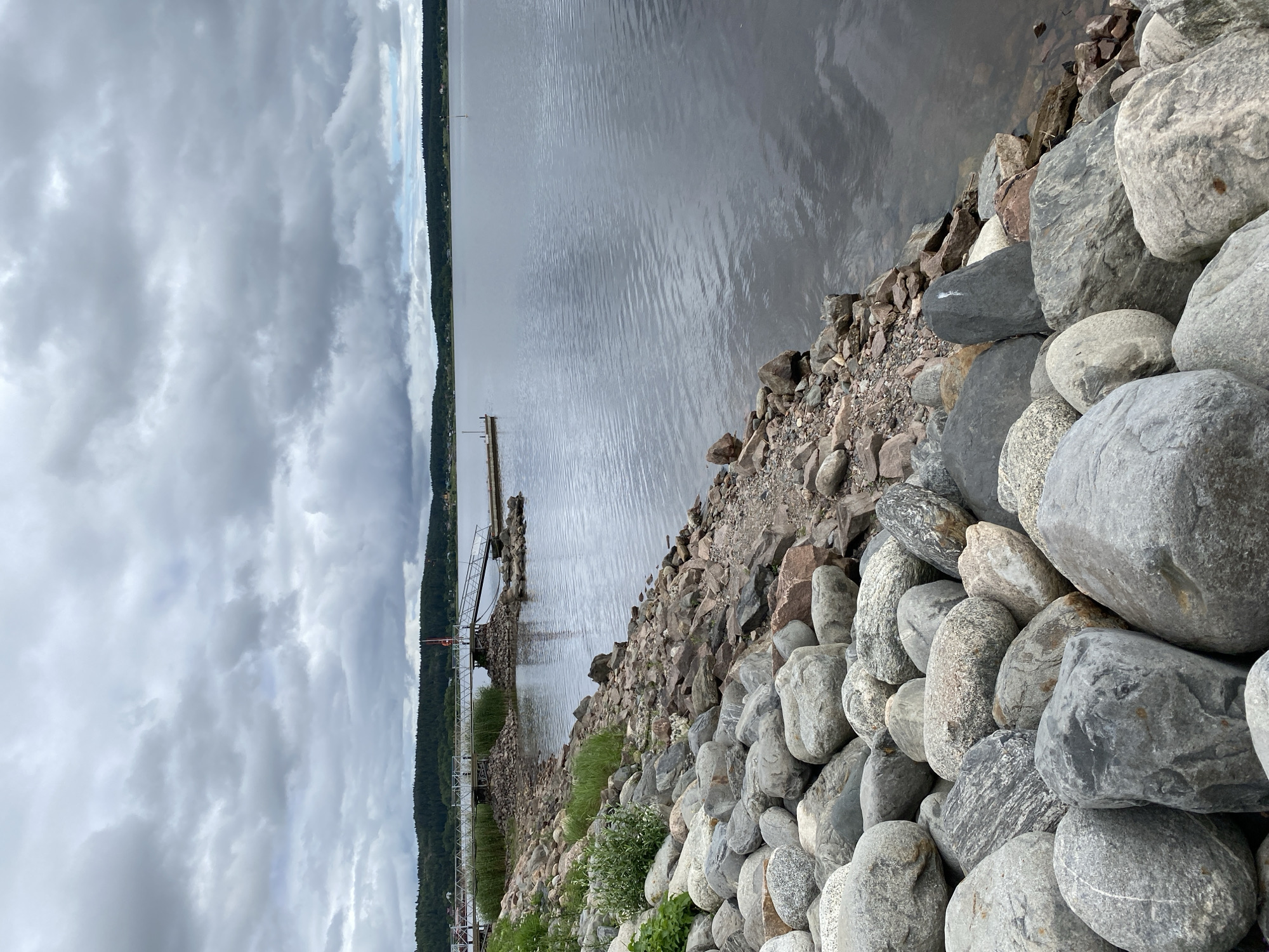 The image depicts a rocky shoreline on a cloudy day, with a calm lake or bay stretching into the distance. A metal pier with a ramp extends from the land into the water. The shoreline is densely covered with a variety of large, rounded stones. Beyond the shoreline, a line of trees forms the horizon. The water's surface is rippled, reflecting the overcast sky. In the far distance, a small sailboat is visible on the water. The overall atmosphere is serene and natural.
