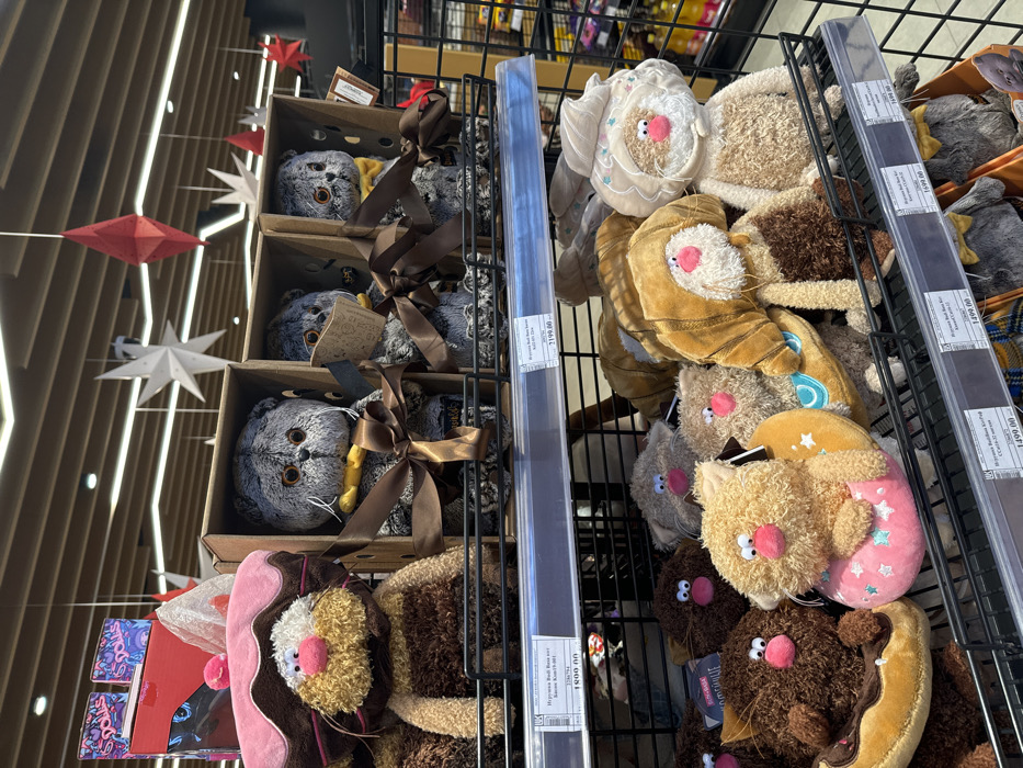 A shelf is filled with stuffed animal toys. Gray stuffed cat toys are displayed in wooden boxes adorned with brown ribbons. Adjacent to the cat toys are stuffed cat toys designed to resemble various foods like donuts and croissants. These are arranged in rows on the shelves. Above the display are more rows of shelves. In the background, decorative stars and diamonds hang from the ceiling.