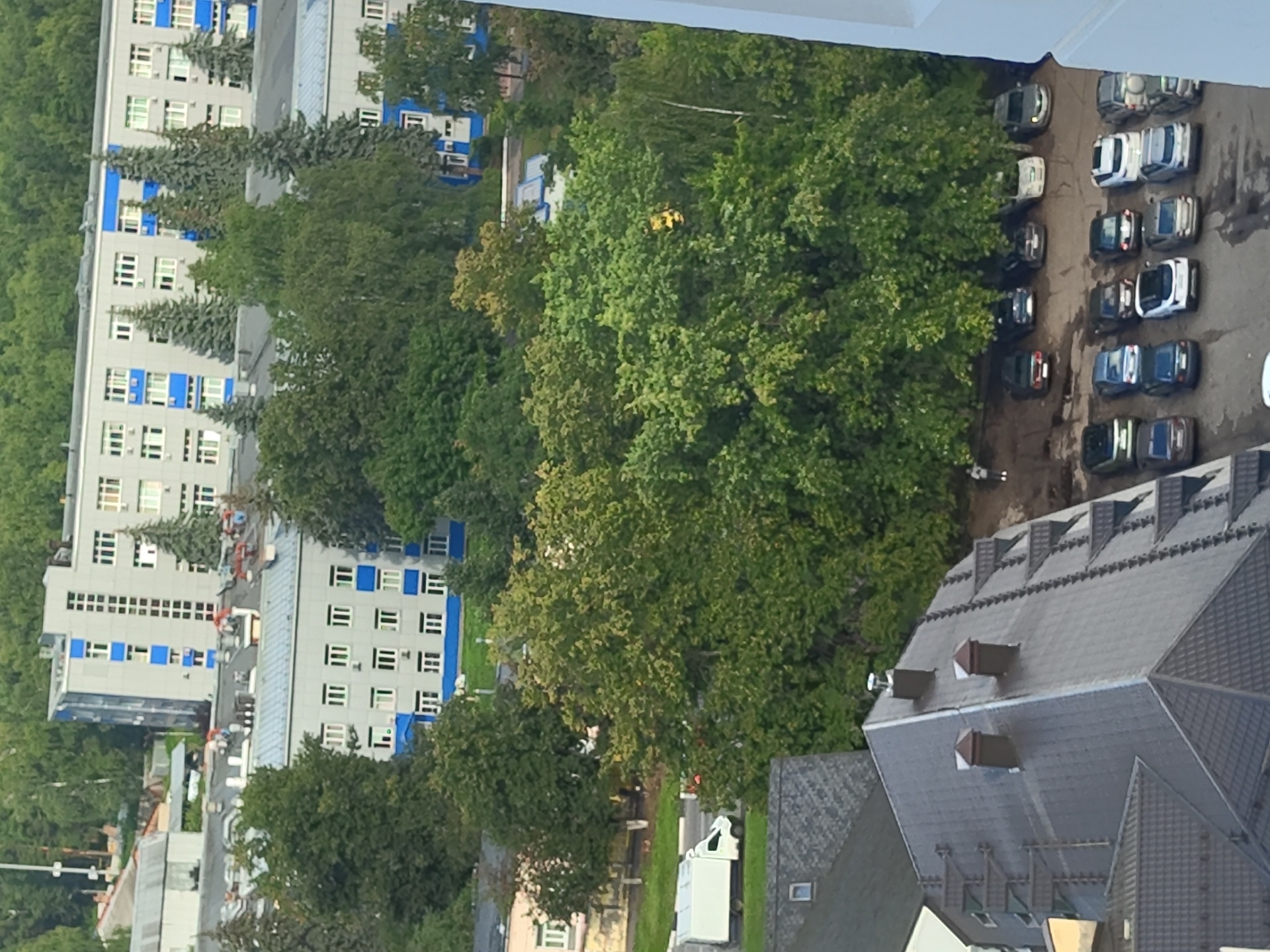 The image depicts an aerial view of a residential area in Ramenskoye, Russia. The scene features several multi-story buildings with gray facades and blue accents, typical of Soviet-era architecture. Large, mature trees with lush green foliage dominate the foreground and middle ground, obscuring parts of the buildings.
In the foreground, a dark gray tiled roof of a building is visible, with several chimneys and what appear to be roof-mounted antennas or supports. To the right of this roof, a parking lot is visible, filled with numerous parked cars of various models and colors, including sedans and SUVs. The parking lot appears to be a dirt and gravel surface. A lone figure, possibly a pedestrian, is standing on the edge of the parking lot near the trees.
Further back, a white delivery truck is driving on a paved road that runs alongside a line of trees. Beyond the trees, more buildings are visible, including a lighter colored building with prominent windows and blue trim. In the distance, a larger, more modern building with blue and gray elements stands out, surrounded by trees. The overall impression is a dense urban environment with significant greenery. The sky is not clearly visible due to the trees and buildings.
