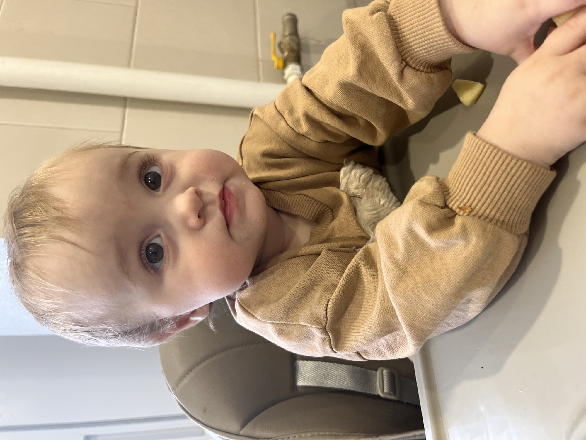 A young child with fair skin and blonde hair, dressed in a tan long-sleeved shirt, is seated in a high chair. The child is looking directly at the camera. A piece of food is on the tray in front of the child. A valve or faucet fixture is visible in the background. The location is Chaikovsky, Russia.