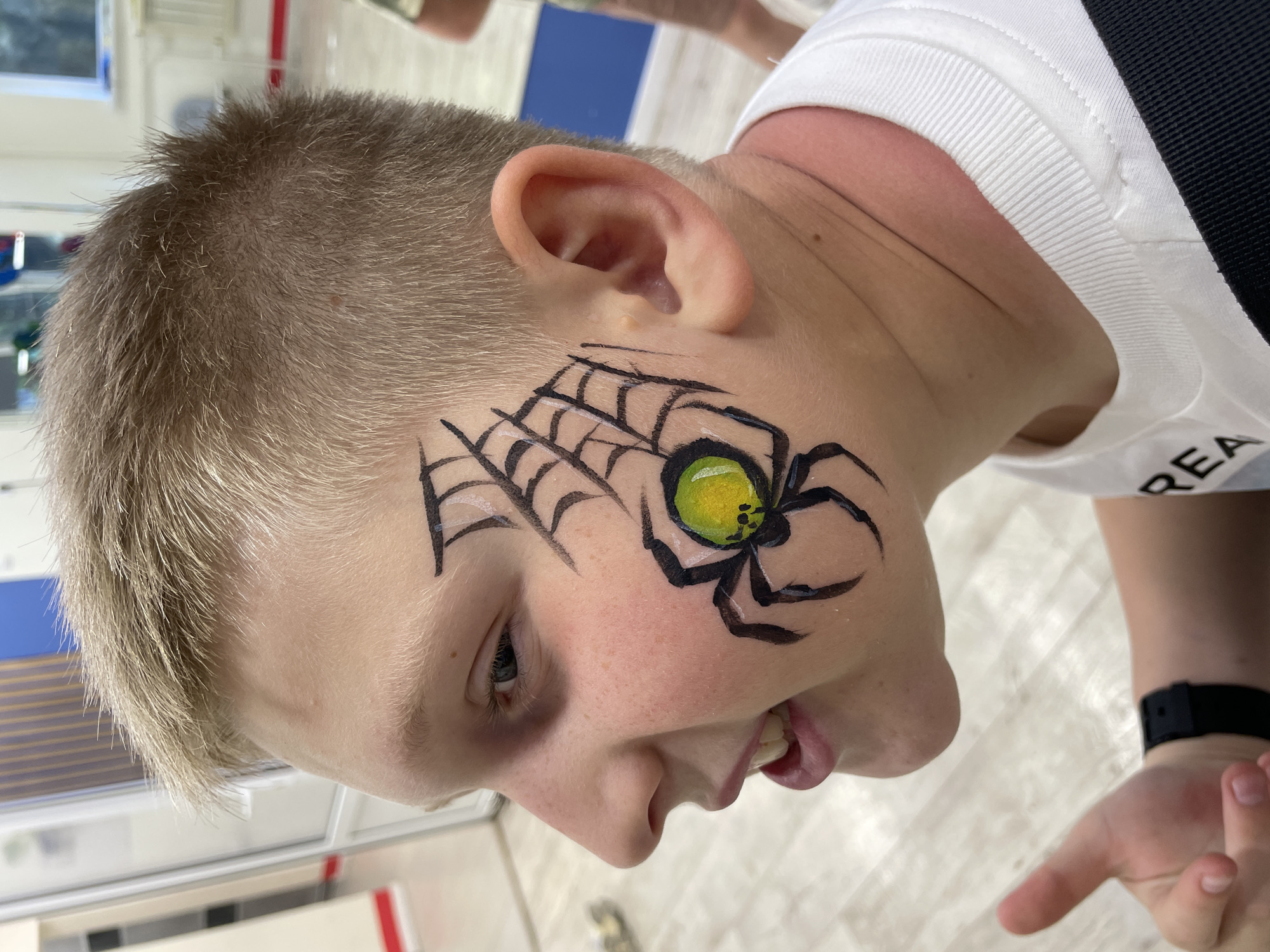 A young boy with short, light blonde or light brown hair and fair skin is depicted in a close-up, angled shot, likely indoors in Sochi, Russia. His right cheek and temple are adorned with vibrant face paint. The design features a detailed black spiderweb with subtle white highlights, stretching from his temple towards his eye. A prominent black spider with a distinctive bright yellow-green body, marked with small black dots, is painted on his cheek below the web. The spider's body appears glossy or reflective.
The boy is wearing a white short-sleeved t-shirt with a ribbed collar or sleeve. A partial word, "REA," is visible in black lettering on his shirt, and a black strap, possibly from a bag or garment, is visible over his shoulder. A black wristband or watch is seen on his left wrist. His mouth is slightly open, revealing his teeth, suggesting he might be smiling, laughing, or mid-sentence, conveying a sense of happiness or excitement.
The background is softly blurred but appears to be a brightly lit indoor space with light-colored walls and flooring, possibly tiles or polished concrete. Hints of blue and red accents are visible in the background, possibly from furniture or decorative elements. The overall impression is a cheerful, lively atmosphere, possibly at a party or event where face painting is offered.