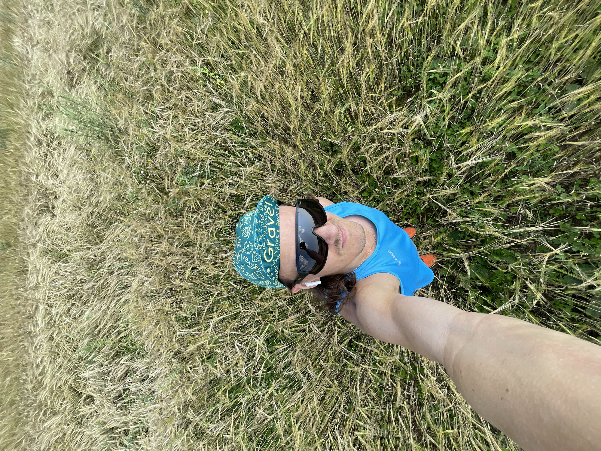 A man is taking a selfie while standing in a field of dry grass. He is wearing a blue tank top with the Jako logo, orange shorts, a green bandana with the word "Gravel" on it, and dark sunglasses. He also has white earbuds in his ears. The field appears to be in Maksimovo, Russia. The image is taken from a high angle, looking down on the man.