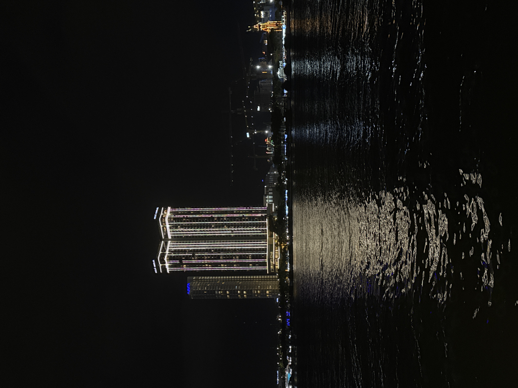 The image shows the Han River waterfront in Da Nang, Vietnam at night. In the foreground, dark water reflects the lights of the city. Dominating the mid-ground are two brightly lit, modern high-rise buildings that appear to be hotels or condominiums. The left building has "AZURA" visible in blue lettering near its top. The other, taller building is illuminated with vertical strips of white, pink, and purple light. Beyond these prominent structures, the city skyline continues with other illuminated buildings, construction cranes, and what appears to be a Ferris wheel or a similar illuminated attraction on the right. The overall scene conveys a vibrant and lively urban nighttime atmosphere.