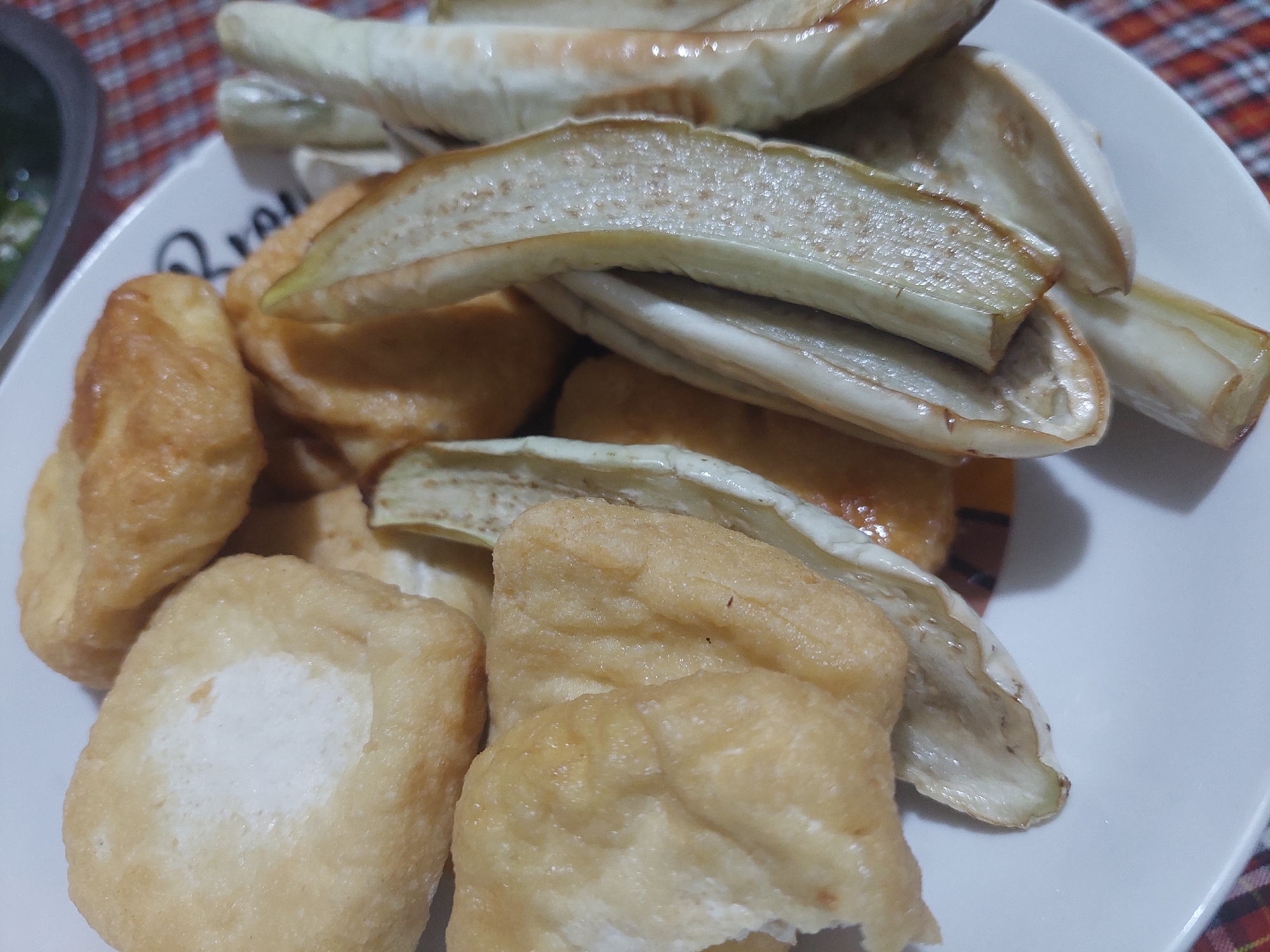 The image shows a close-up of a white plate filled with food. On the left side of the plate are several pieces of fried tofu, which are golden brown and appear fluffy. In the center and right side of the plate are strips of cooked eggplant. The eggplant is light green and white, with some darker speckles, and has a slightly glossy appearance, suggesting it has been cooked in oil. The food is arranged on a white plate with a black rim that has some text on it, partially obscured. The plate sits on a red and white checkered tablecloth. In the upper right corner, out of focus, a portion of a clear plastic container with some green food items is visible. The image does not provide any specific information or context that would allow for the determination of its location in Jelutung, Indonesia.