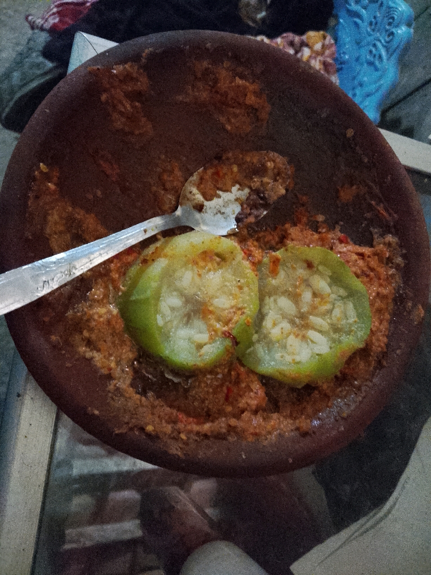 A close-up shot shows a traditional Indonesian mortar and pestle, made of dark clay, filled with a reddish-brown sambal, a spicy chili paste. Two slices of green fruit, likely a type of guava or young papaya, are placed on top of the sambal. A metal spoon is resting partially in the mortar, its handle extending out of the frame. The mortar and its contents are set on a reflective surface, possibly a glass table, which shows a blurred reflection of the surroundings. In the background, a bright blue decorative object and a few other items are out of focus, suggesting an indoor setting, possibly a home kitchen in Wadang, Indonesia, where the meal is being prepared or served.