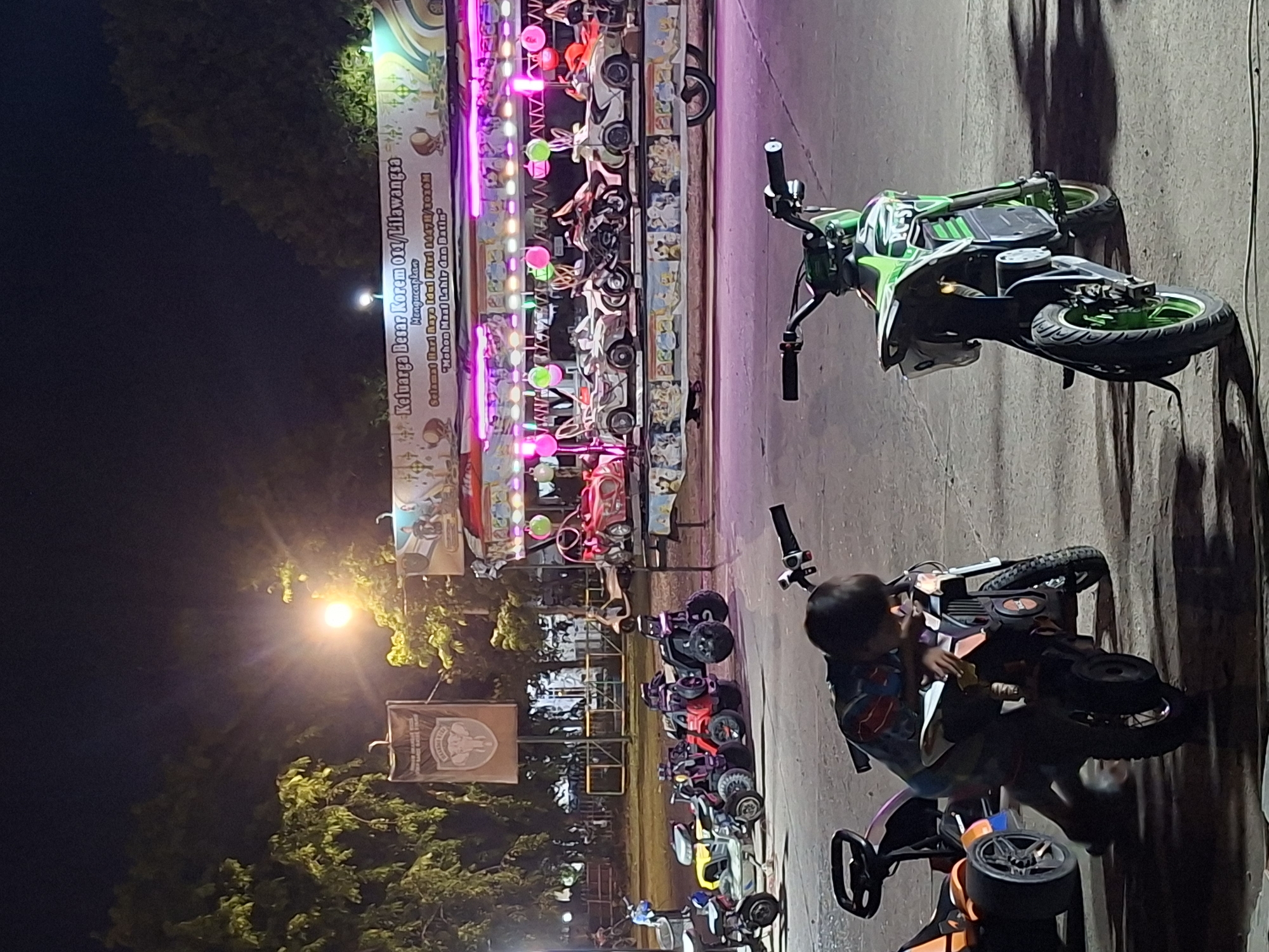 This image captures a vibrant night scene at an outdoor entertainment venue, possibly a fairground or recreational area, in Lhokseumawe, Indonesia. The setting appears to be a paved or concrete area with a dark sky, illuminated by numerous artificial lights.
In the foreground, a child with dark hair is visible, partially obscured, appearing to interact with or sit on a small orange and black ride-on vehicle, possibly a mini ATV. Nearby, a green and black toy motorcycle stands upright. Further into the scene, a variety of other colorful ride-on vehicles, including miniature motorcycles, ATVs, and cars in red, yellow, and black, are lined up or scattered, suggesting a rental or play area.
These vehicles and the surrounding structures are brightly lit with string lights and neon-like fixtures, predominantly in shades of pink, purple, and green, creating a festive atmosphere. On the far left, a tall banner or sign is prominently displayed and well-lit. The text on this banner, as deciphered by OCR, reads: "Keluarga Besar Korem 011/Lilawangsa Mengucapkan Selamat Hari Raya Idul Fitri 1447H/1936M 'Mohon Maaf Lahir dan Batin'". This indicates an Eid al-Fitr greeting from a military unit.
A bright street lamp casts a strong glare on the left side of the image, and trees are visible in the background, silhouetted against the night sky. The overall scene depicts a lively recreational event, likely catering to children, taking place during the Eid al-Fitr celebrations.