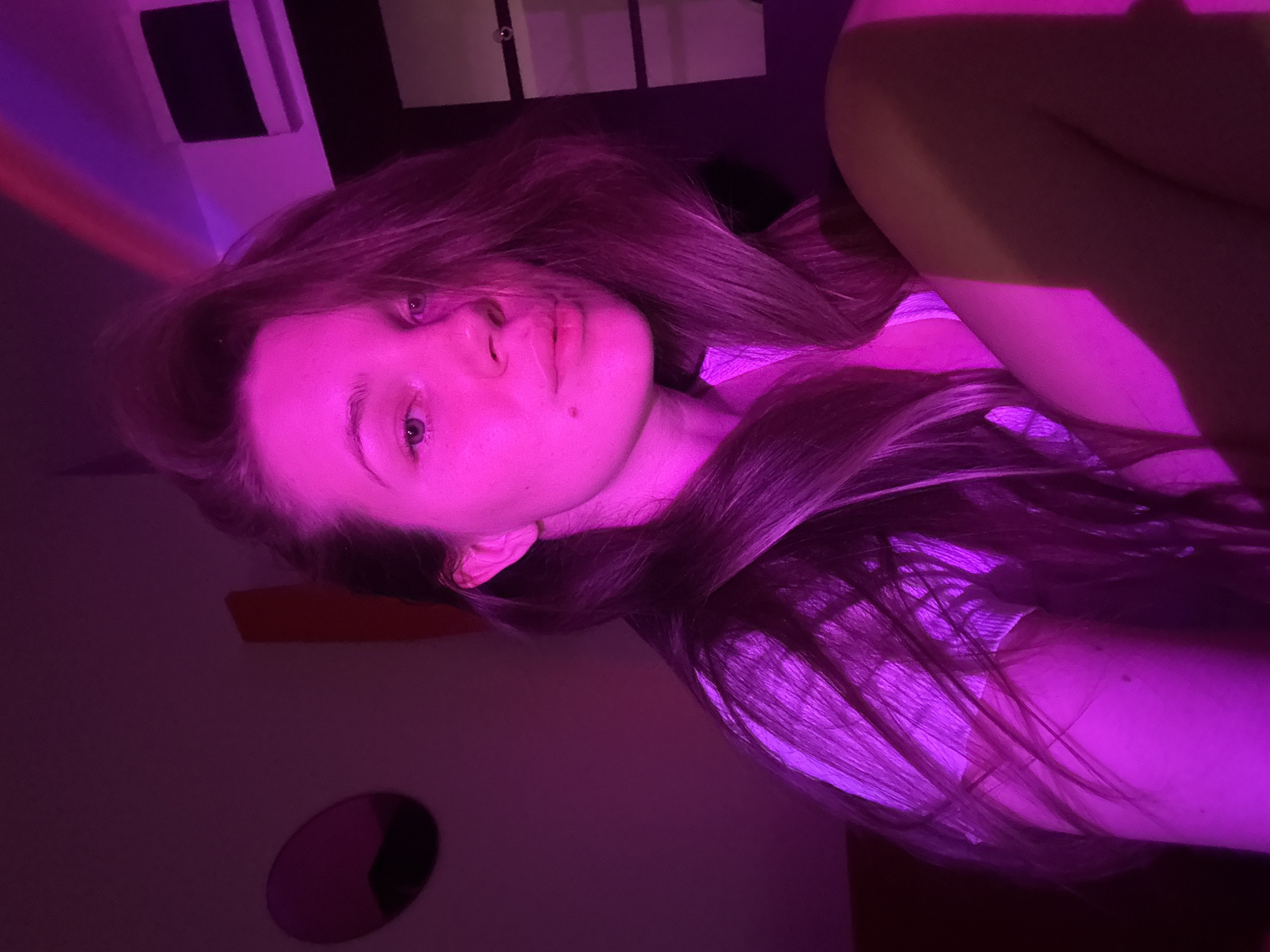 A dimly lit indoor scene, bathed in intense pink and purple lighting, features a Caucasian woman. The lighting obscures details, but the woman has light skin, light-colored hair, and is wearing what appears to be a white or light-colored top. She is positioned at an angle to the camera, her face the primary focal point. In the background are indistinct shapes, possibly including a window, a mirror, and wall fixtures. The location, Vozdovac Urban Municipality, Serbia, cannot be verified from the image alone.