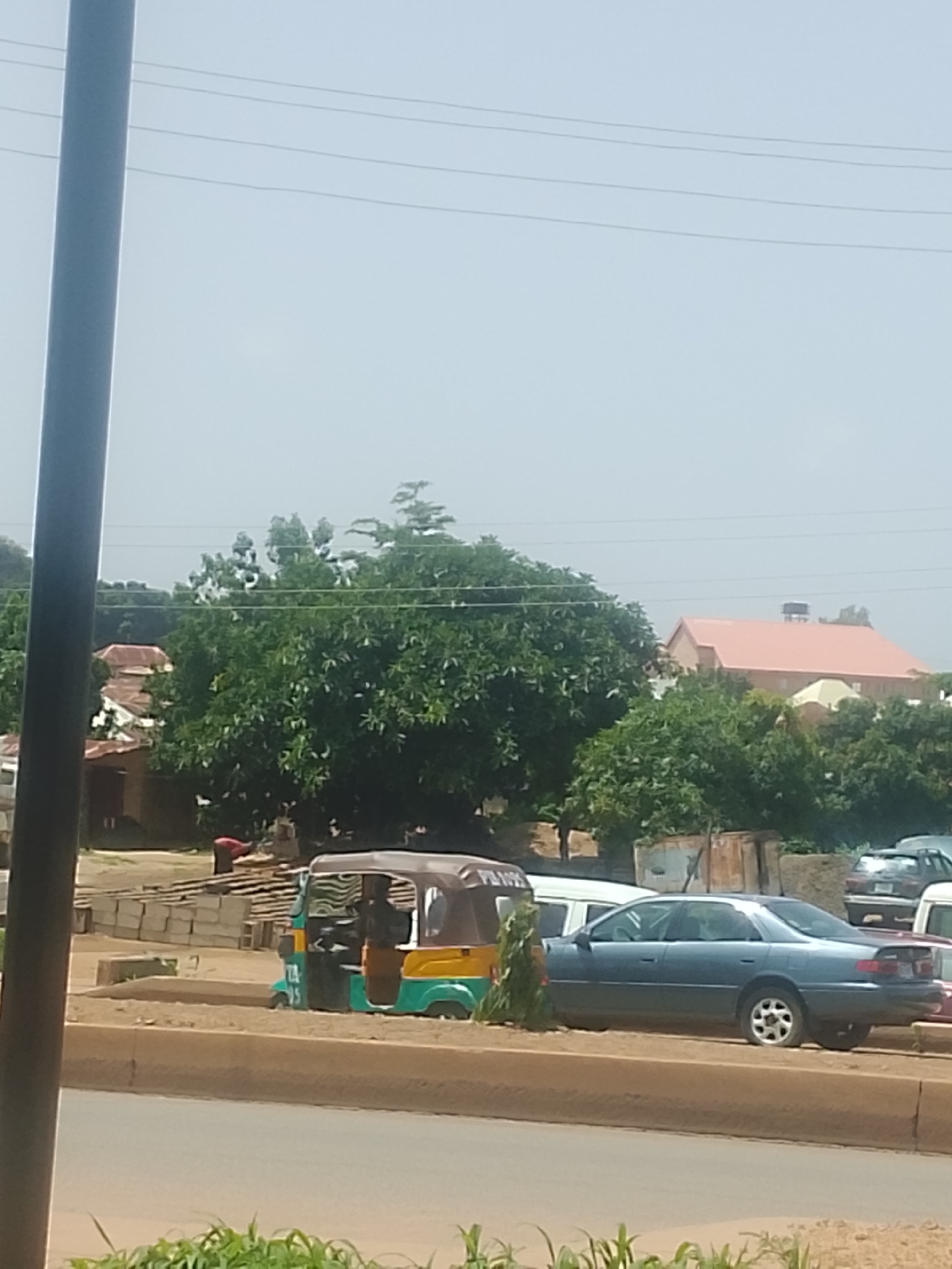 This image captures a street scene in Bukuru, Nigeria. A light blue sedan is parked prominently on the right side of the frame, facing left. To its left, a colorful auto-rickshaw, predominantly green and yellow, is also parked. The auto-rickshaw has a driver visible inside. In the background, several buildings with reddish roofs and lush green trees are visible, suggesting a warm, sunny day. Power lines are strung across the sky. To the left of the auto-rickshaw, a pile of what appears to be bricks or construction materials is stacked on the side of the road. A person in a red shirt is bent over near these materials, possibly working. The overall scene depicts a typical day in a Nigerian town with a mix of vehicles and everyday activities. The time of day appears to be daytime, given the bright lighting and clear sky.