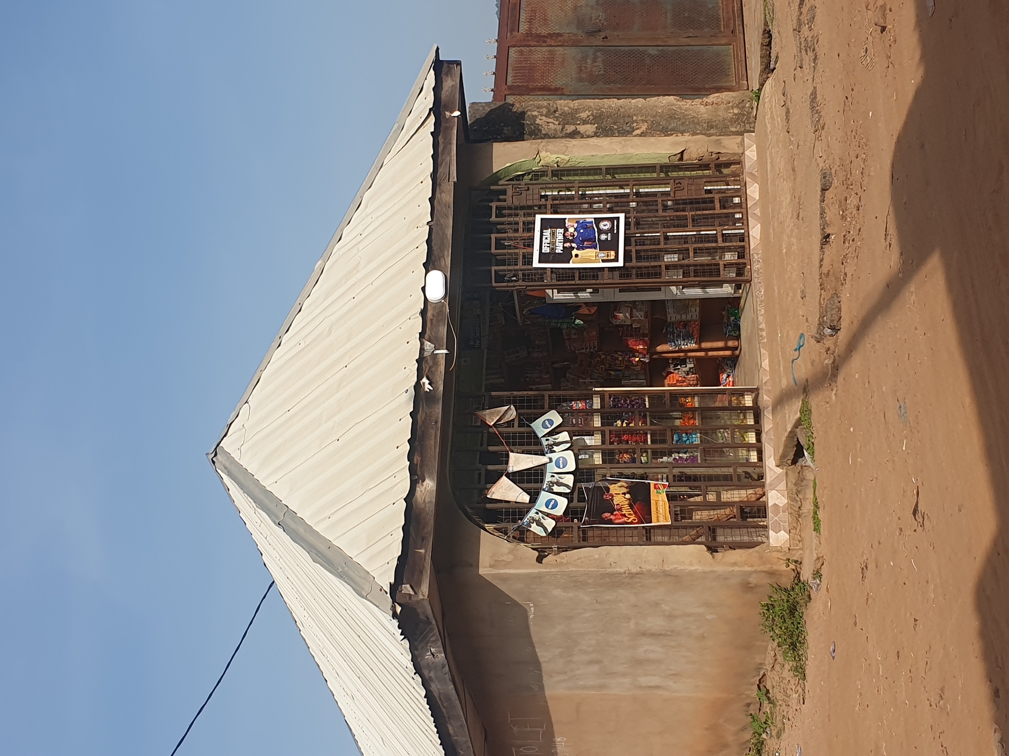 This is a medium shot of an outdoor market stall with a corrugated iron roof, illuminated by the bright sun, suggesting midday. The scene is set in Lokoja, Nigeria, indicated by the context provided. The stall is built into a wall, with metal bars securing the entrance, revealing shelves stocked with various goods.
In the foreground, a dark shadow stretches across a dusty, uneven ground, hinting at the position of the sun. The sky above is a clear, bright blue, characteristic of a sunny day. The stall itself is a triangular structure made of light-colored corrugated metal. A white outdoor light fixture is mounted on the side of the roof.
Attached to the metal bars of the stall entrance are several items. A colorful banner or poster hangs prominently, displaying images of people in sports attire and the text "OFFICIAL PARTNER". Below this, a string of small triangular flags or banners are displayed, each with a blue logo and white text, possibly branding for a product. Further into the stall, other colorful products are visible on the shelves.
To the right of the stall, there's a rusted metal gate above the stall. The overall impression is of a typical street-side market in a warm climate, with a sense of daily commerce taking place under a clear sky.