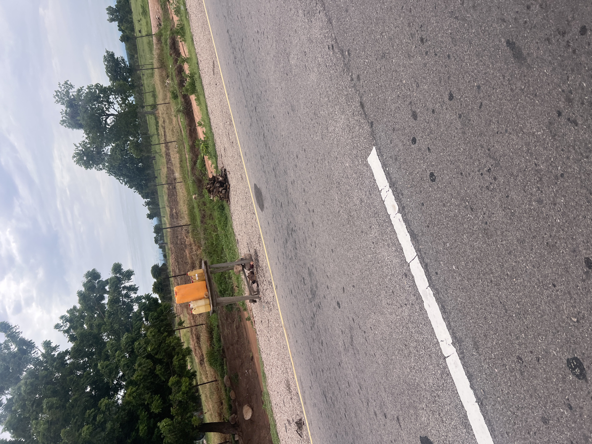The image shows a roadside scene in Minda, Nigeria. The primary subject is a paved road with a white line marking the lane. To the left of the road, there is a gravel shoulder followed by a fence and then a field with scattered trees. In the foreground, there is a wooden stand with three containers on it: one large orange container and two smaller yellow containers. The scene appears to be set during the daytime, with a cloudy sky. The overall impression is of a rural or semi-rural landscape, possibly along a main road in Nigeria. There is no text visible in the image, and no people or specific activity are depicted, other than the general context of a roadside. The image is taken from a slightly elevated perspective, looking down at the road and the adjacent land.