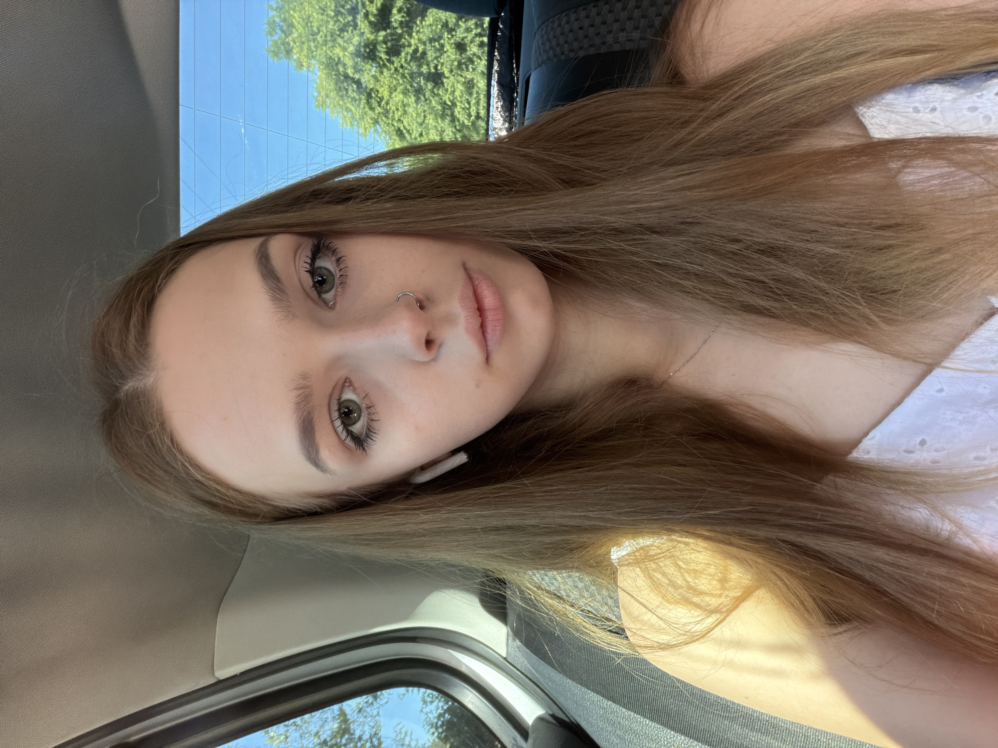 A young woman with long, light brown hair is pictured in a selfie taken from inside a vehicle. She is wearing a white top with eyelet details. She has a silver hoop nose ring and is wearing white earbuds. The background shows the sky and trees through the rear window of the car, suggesting it is daytime and fair weather. Sunlight is casting shadows on the woman and interior of the car. Her expression is neutral, and the overall tone of the image is casual. No visible text is present.