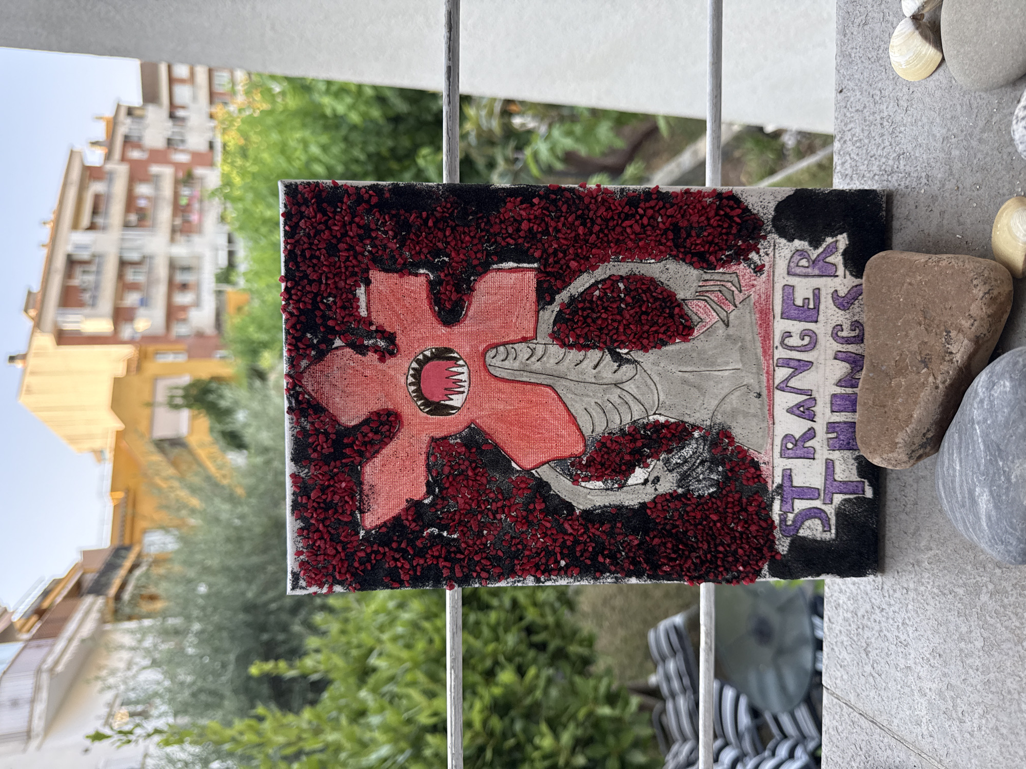 A framed canvas painting depicting the Demogorgon from the TV show Stranger Things sits on a ledge outdoors. The painting features the creature's signature red, flower-like head with sharp teeth and a grey, humanoid body. The background of the painting is a textured collage of small red and black pieces. Below the creature, the words "STRANGER THINGS" are painted in purple letters. The canvas rests on a textured grey surface, possibly a balcony or patio, with several stones and shells scattered in front of it. In the background, out of focus, are buildings and lush green trees, suggesting an urban environment with vegetation. The bright sky and natural light indicate it is daytime. The overall scene conveys an artistic display set against a residential backdrop.