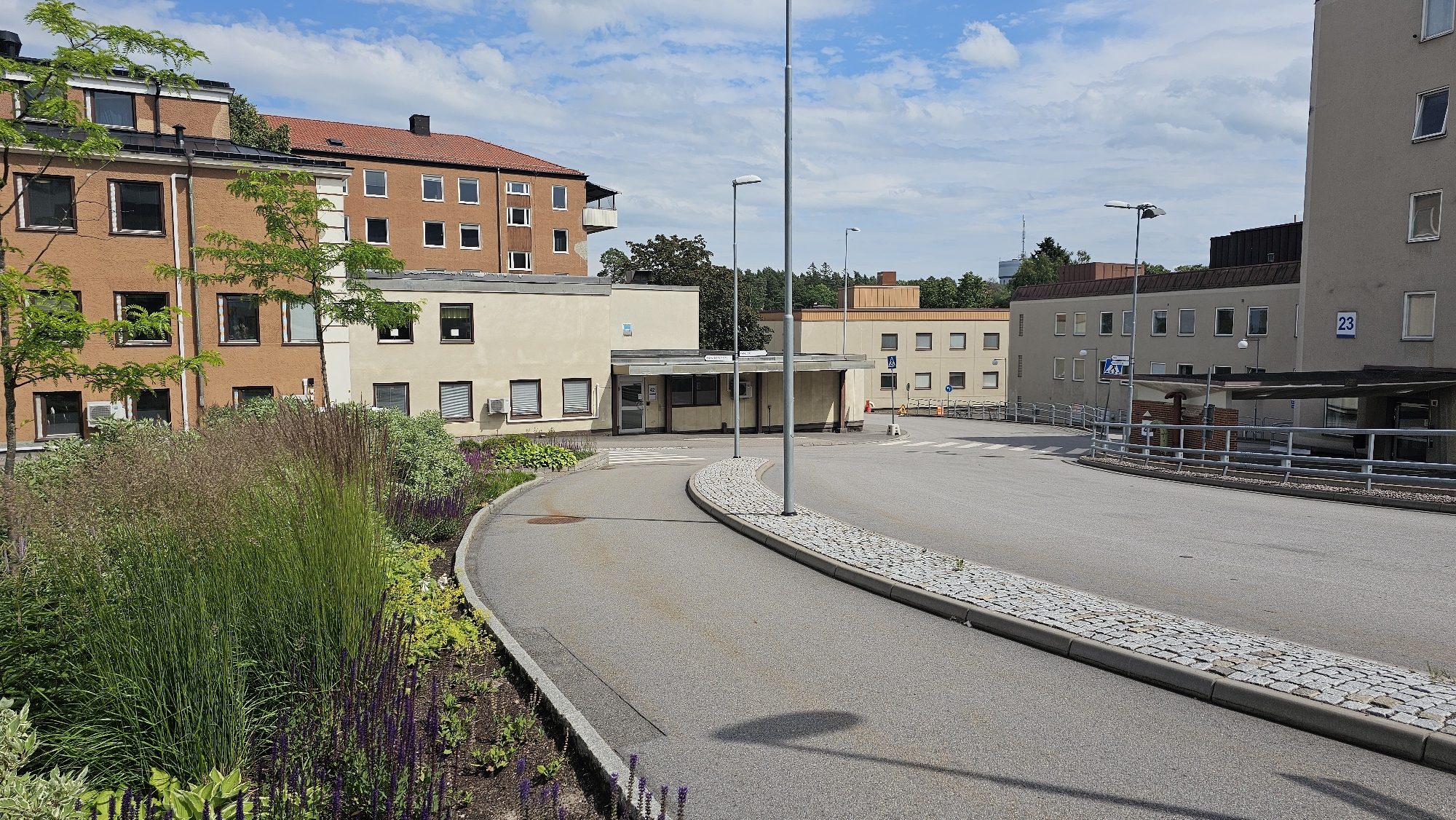 This media file depicts an outdoor scene at a medical facility or hospital in Linköping, Sweden, under a bright, cloudy sky, suggesting it is daytime. The image shows a complex of buildings, likely part of a hospital, with multiple structures of varying heights and architectural styles. The foreground features a gently curving asphalt road leading into the scene, bordered by a meticulously landscaped garden filled with tall grasses and purple flowers.
The buildings are modern, with several featuring beige or light gray facades and numerous rectangular windows. One prominent building on the left has an orange-brown facade with a reddish-brown tiled roof, and a balcony. Another building in the background is also reddish-brown. A smaller, single-story building with a covered entrance and a sign in Swedish is situated near the center of the frame. Various trees and greenery are visible, adding to the pleasant, somewhat tranquil atmosphere of the setting.
Several lampposts are strategically placed along the road and pathways. A blue sign with the number "23" is visible on the side of a building on the right. There is a pedestrian crossing marked on the road. The overall impression is one of a functional and well-maintained healthcare environment, situated within a green and landscaped area. No people are visible in the frame, indicating a quiet moment at the location. The weather appears to be warm and sunny, with scattered clouds casting soft shadows.