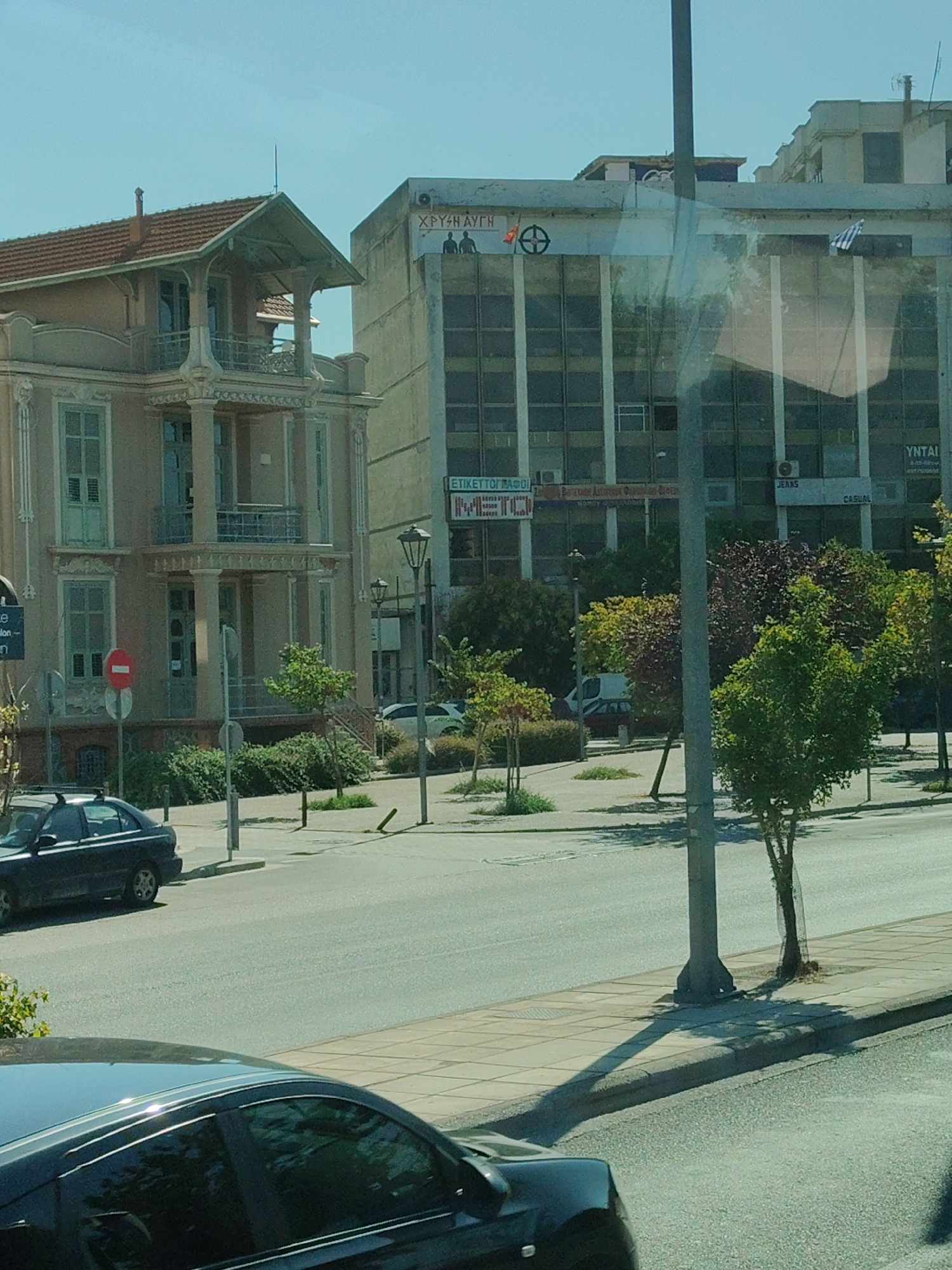 The image captures a street scene in the city of Thessaloniki Municipal Unit, Greece. In the foreground, a dark-colored sedan is parked on the right side of the street, with its roof rack visible. Further down the street, another dark-colored sedan is parked on the left side. A light-colored sedan is visible in the background, also parked on the left.

The environment is urban, with paved roads and sidewalks. A variety of trees and greenery are present along the sidewalks and in front of buildings. Several streetlights and traffic signs are also visible.

Two buildings dominate the background. The building on the left is a pale pink, multi-story structure with ornate architectural details, balconies, and arched windows. The building on the right is a larger, more modern, multi-story building with a glass facade. Signs on this building are visible, including one with Greek lettering and a Greek flag attached. One sign appears to indicate a shop selling "JEANS" and "CASUAL" wear. Another sign features Greek lettering and what appears to be a symbol.

The sky is clear and blue. The overall impression is that of a sunny day in a Greek city.