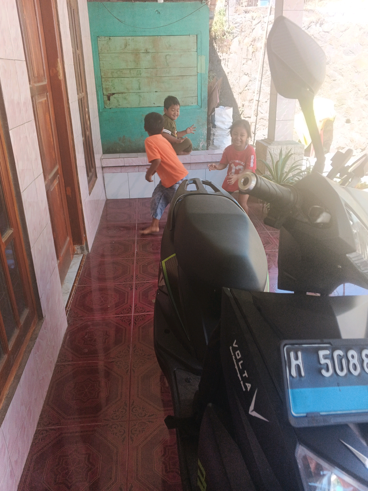 In the city of Keji, Indonesia, on a sunny day, three children are playing outside a building. A young boy in an orange t-shirt and blue pants runs across a tiled floor, his bare feet visible. Next to him, another boy in a green t-shirt sits on a low wall, smiling. To the right, a young girl in a red t-shirt laughs as she stands near a potted plant. In the foreground, a black motorcycle is parked, its license plate reading "H 5088". The background features a rocky wall and a blue wooden door.