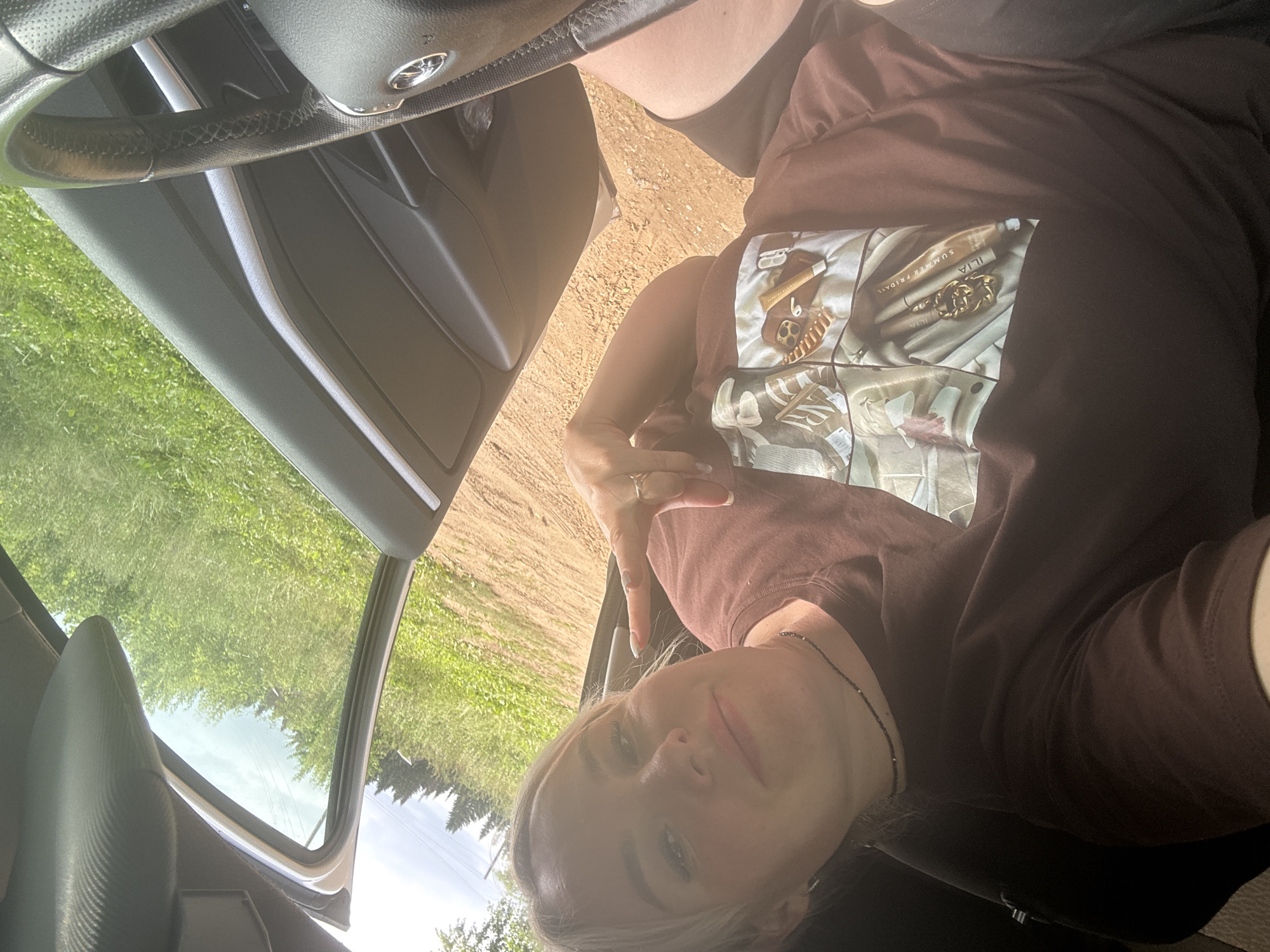 A woman with blonde hair and makeup is taking a selfie inside a car. She is wearing a brown t-shirt with a graphic print on the front and a black beaded necklace. Her left hand is raised, and she is making a gesture with two fingers. The car door is open, revealing a grassy area with trees in the background. The location is the city of Nozhovka, Russia. The scene is illuminated by daylight.