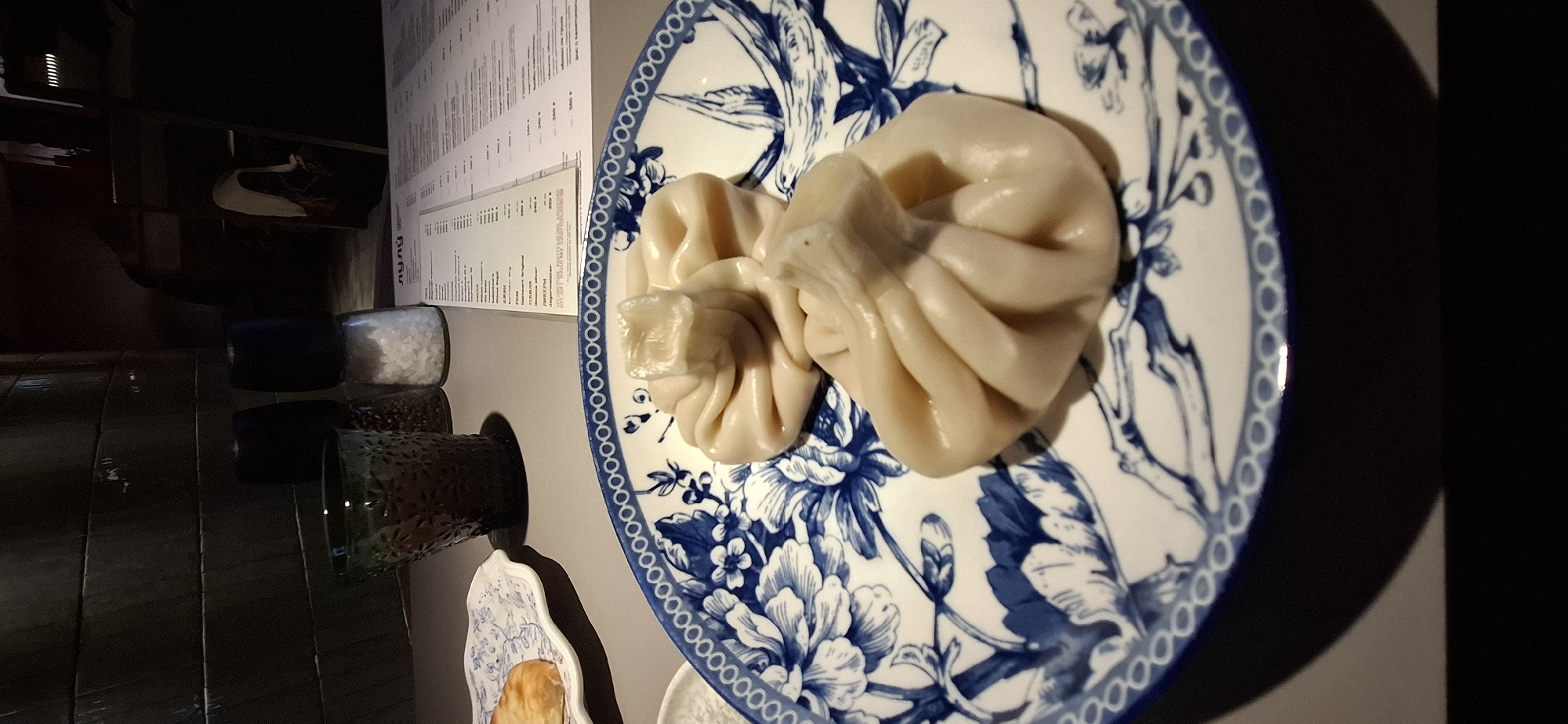 The image shows two khinkali on a blue and white patterned plate, likely taken in a restaurant. The plate is on a gray surface. To the left, there are condiments in glass containers, and a menu board is visible in the background. The location is presumably in Chaikovsky, Russia, based on the context.
