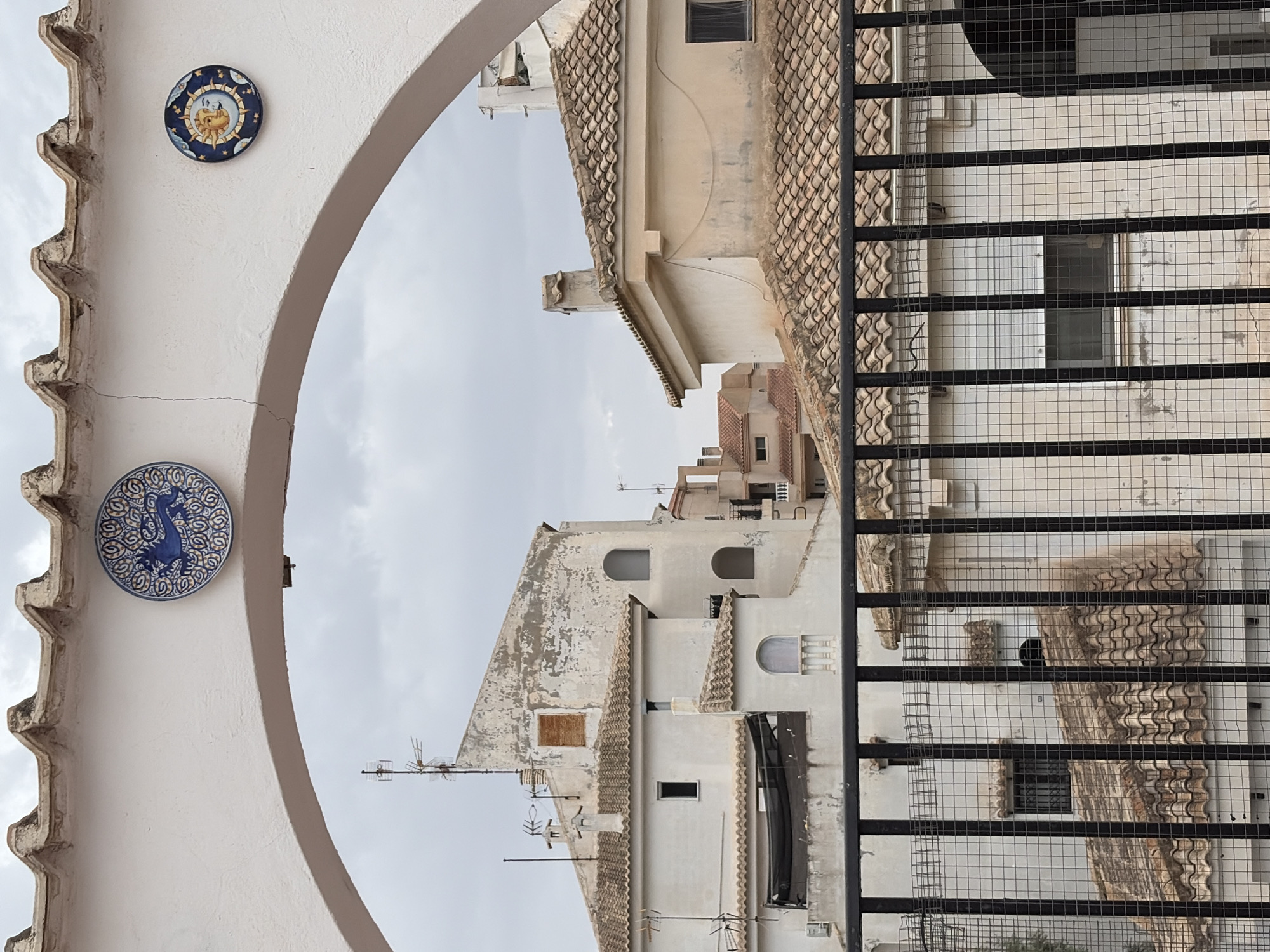 The image shows a view through a white arched structure with decorative ceramic tiles mounted on its surface, looking out onto a cityscape under an overcast sky. The left tile depicts a blue lion or mythical creature with intricate patterns, and the right tile features a sun and moon face. In the background, white and beige buildings with terracotta tiled roofs are visible. Some buildings appear aged with peeling paint. A black metal fence with a wire mesh overlay is in the foreground, obscuring parts of the lower buildings. Antennae and chimneys are present on several rooftops, indicating a residential or urban area. The scene is likely from an elevated vantage point, possibly a balcony or rooftop, in Torrevieja, Spain.