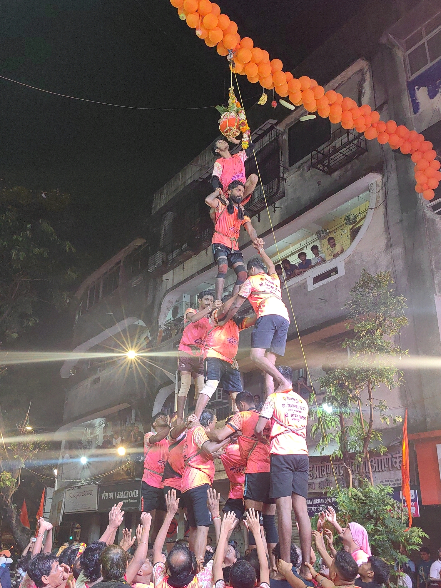 This image captures a scene from a Dahi Handi festival event, likely in Mumbai, India, during the nighttime. A human pyramid is being formed by a group of men dressed in vibrant orange and pink t-shirts, with some wearing black shorts. They are stacked several layers high, with the bottom layer of men standing on the ground, forming a base. The men in the pyramid are supporting each other, with some holding hands and others using their shoulders and backs to create stability. At the very top of the pyramid, a single individual is reaching towards a pot suspended by a rope, decorated with flowers and possibly containing curd (dahi) or butter. The event is taking place outdoors, with a building visible in the background, featuring balconies and windows where spectators can be seen watching the proceedings. The area is adorned with a garland of orange balloons, adding a festive atmosphere to the occasion. Several people in the foreground are looking up, with their hands raised, likely cheering and encouraging the participants in the pyramid. The lighting suggests a public gathering, with artificial lights illuminating the scene and creating a warm glow.
