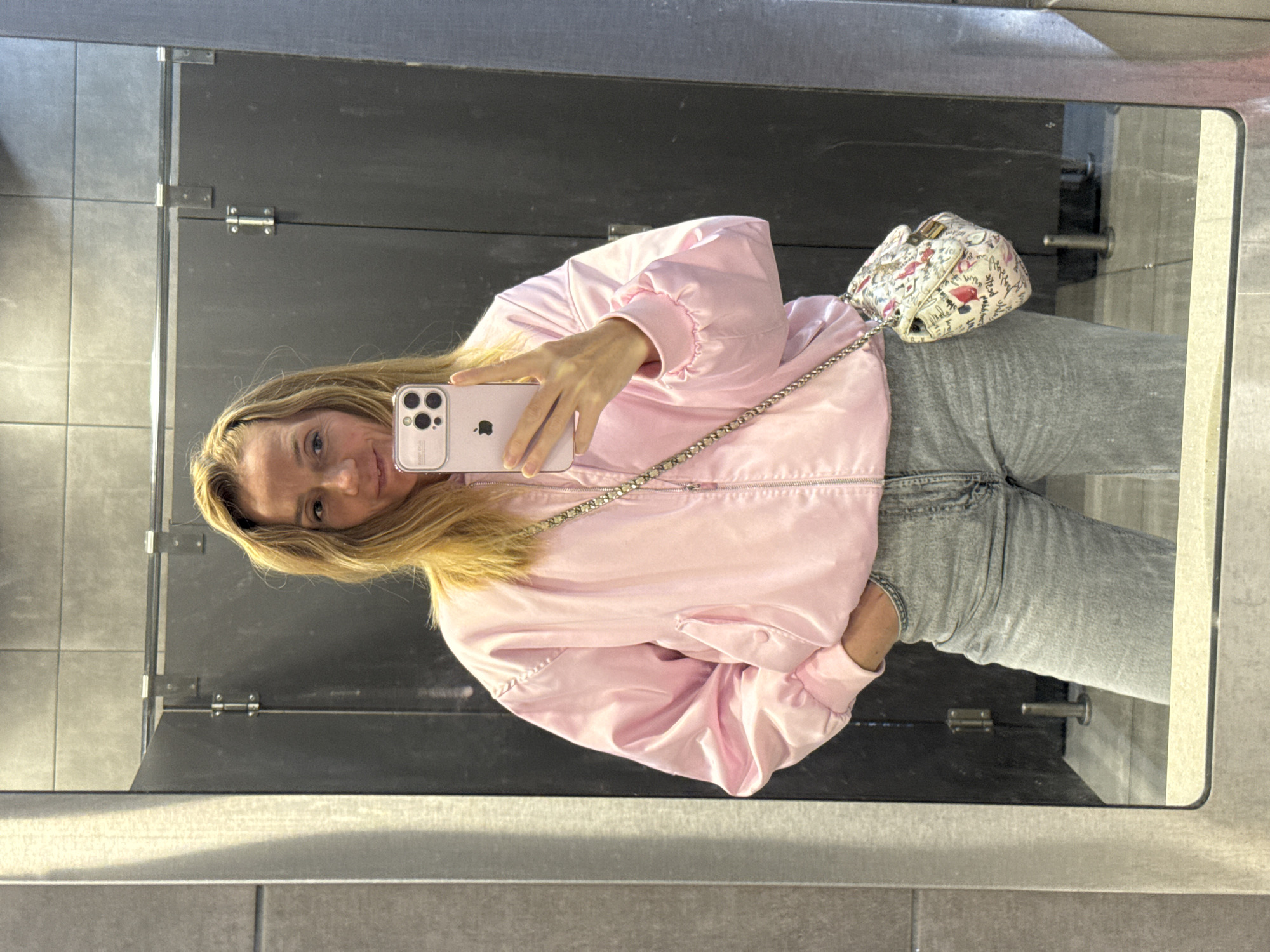 A blonde woman is taking a selfie in what appears to be a public restroom. She is wearing a pink satin jacket, gray jeans, and a small white patterned purse with a chain strap. She is holding a light pink iPhone in her right hand, angled to capture her reflection in the mirror. The background features gray stall dividers with metal hardware. The restroom has gray tiled walls. There is no indication of the location being in Saint Petersburg, Russia.