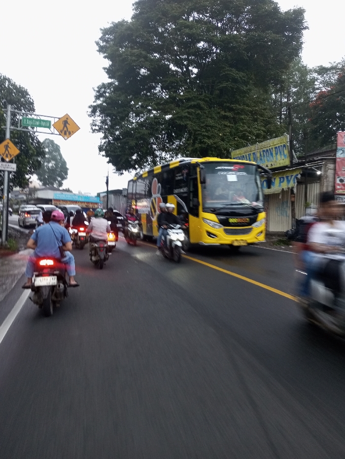 The image displays a traffic scene on a wet asphalt road in Dawathang_Dorjibi_ Kashingtsawa, Bhutan, under an overcast sky. The road features a solid yellow center line and a white shoulder line on the left.
Multiple vehicles are present. In the foreground and mid-ground, numerous motorcycles are navigating the roadway. The closest visible motorcycle, a dark-colored scooter, has a rider in a blue shirt and a pink cap, and a passenger, both wearing sandals. The scooter's taillight is illuminated, and its rear license plate reads "F 5391 KP". Other motorcycle riders are visible behind, some wearing helmets and one in a white shirt.
A large, modern yellow bus with black accents and "JAVANEST" branding is prominent on the right side of the road, facing the viewer's left. It appears to be in an adjacent lane or part of slow-moving traffic. Its front is distinctly visible with large headlights. Further down the road, additional cars are visible in the distance on the left.
Roadside features include a green street sign with white text reading "Jl. Raya Ciapus - Parakan" on the left, mounted below a yellow diamond-shaped pedestrian crossing sign. Another identical pedestrian sign is visible further along the road. The upper background is dominated by dense green foliage from large trees. On the right, behind the bus, a commercial structure with a corrugated metal facade displays a sign advertising "JUAL PLAFON PVC". Traffic appears to be moving slowly.