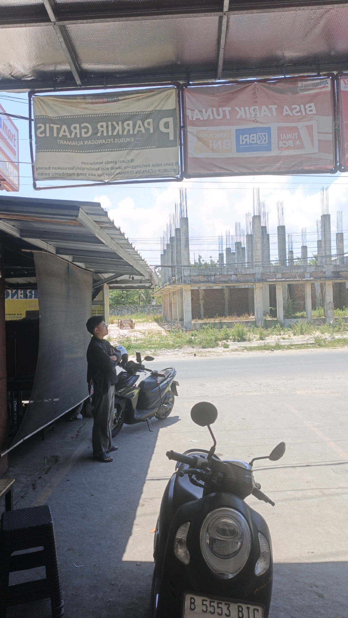 A medium shot shows a man in a black shirt and grey pants standing with his arms crossed, facing a construction site in the distance. The man is positioned under a covered walkway with a large, dark grey tarp hanging to his left. To his right are two parked motorbikes, one black and one dark colored. The construction site features unfinished concrete pillars and brickwork. Above the man and the walkway, banners are visible. One banner, predominantly white and green, reads "GRATIS PARKIR" in large letters, indicating free parking. Another banner, red and white, advertises banking services with logos for "BNI" and "BCA". The scene is set outdoors on a sunny day, with a paved road in the foreground. The location is identified as Desa Pagandaran, Indonesia.