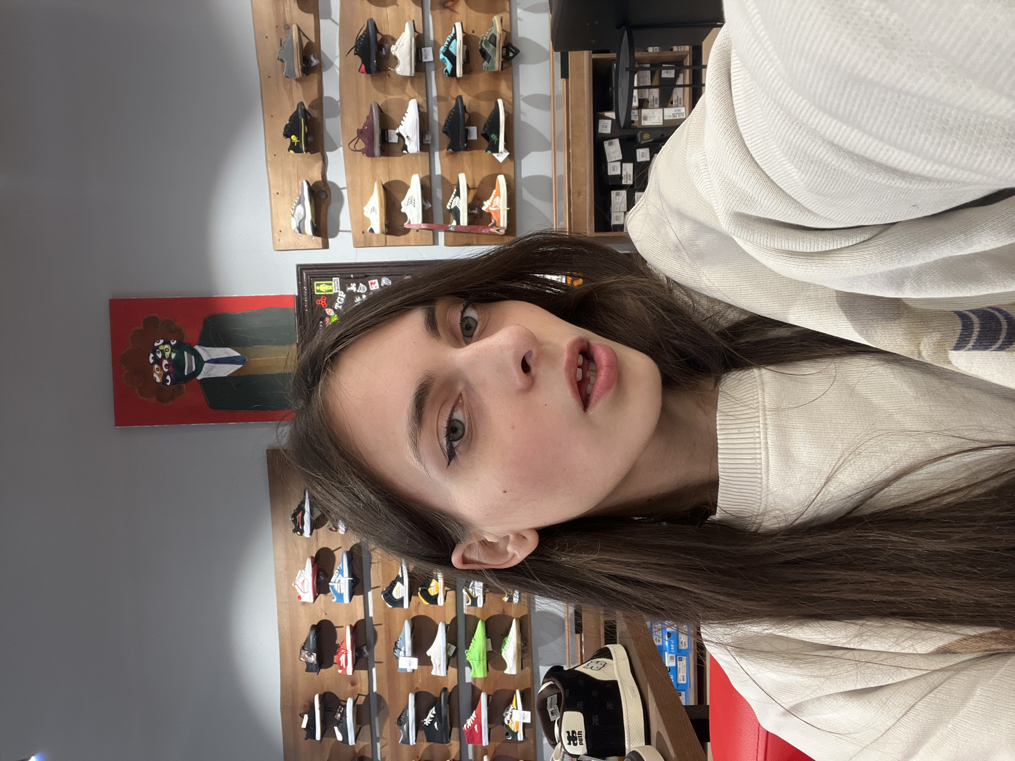 A young woman is facing the camera at a tilt. She has fair skin, brown hair, and green eyes. She is wearing a light-colored sweater. Behind her is a wall display of shoes. There is also a framed picture on the wall. The picture shows an individual with a brown afro, wearing a black suit. Based on the shoe selection, this appears to be a shoe store. Given the presence of Cyrillic characters on a box behind her and the location information provided, the location is likely a shoe store in Krasnoyarsk, Russia.