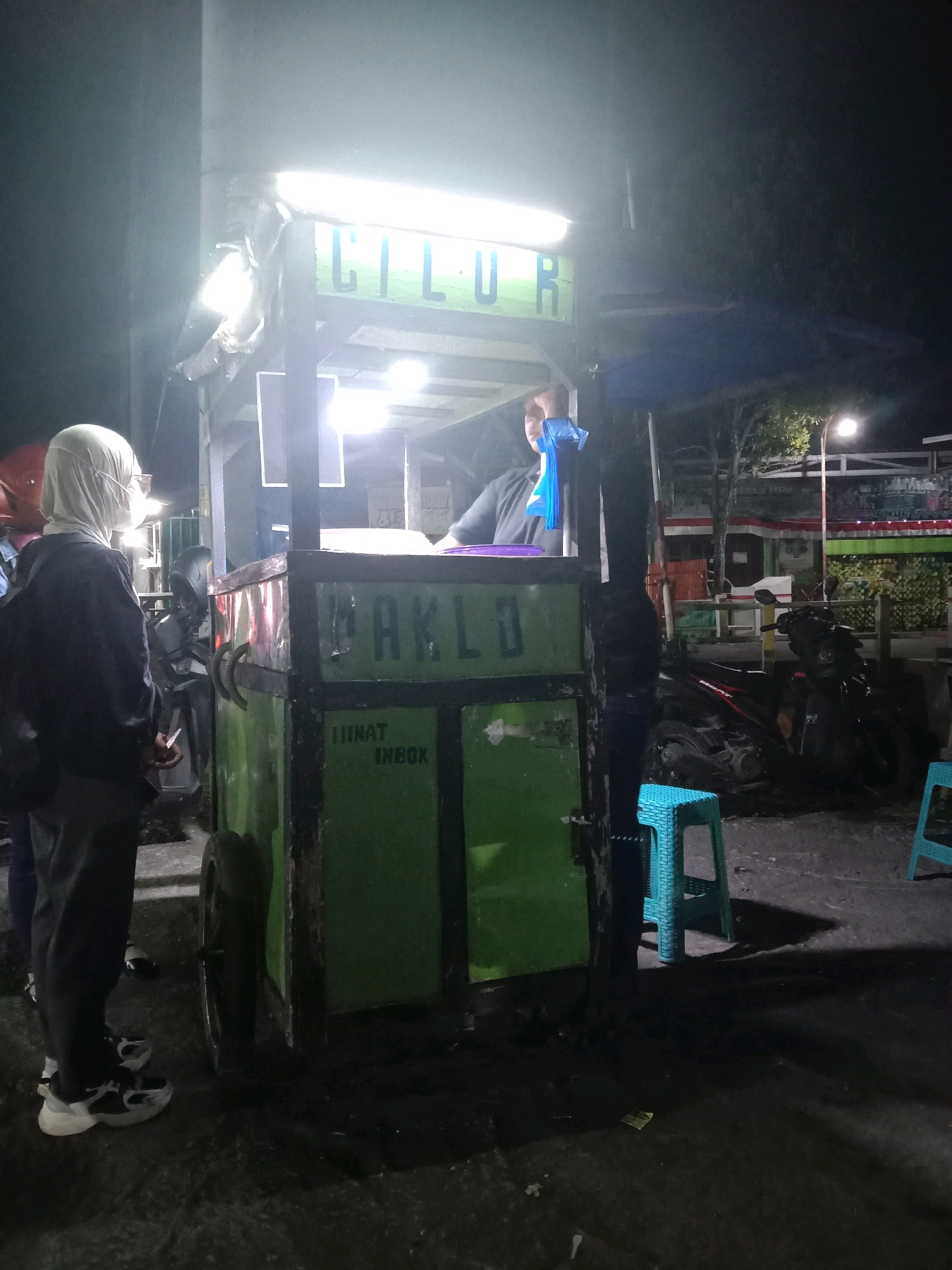 A street food cart with "CILUR" and "MAKLO" written on it is illuminated at night. A person wearing a white headscarf and mask stands to the left of the cart, facing away from the camera. Another person, likely the vendor, is partially visible behind the cart. Several motorcycles are parked behind the cart, and two teal stools are placed on the ground in front of it. The scene appears to be in a public area in Kebonsari, Indonesia, as suggested by the context.