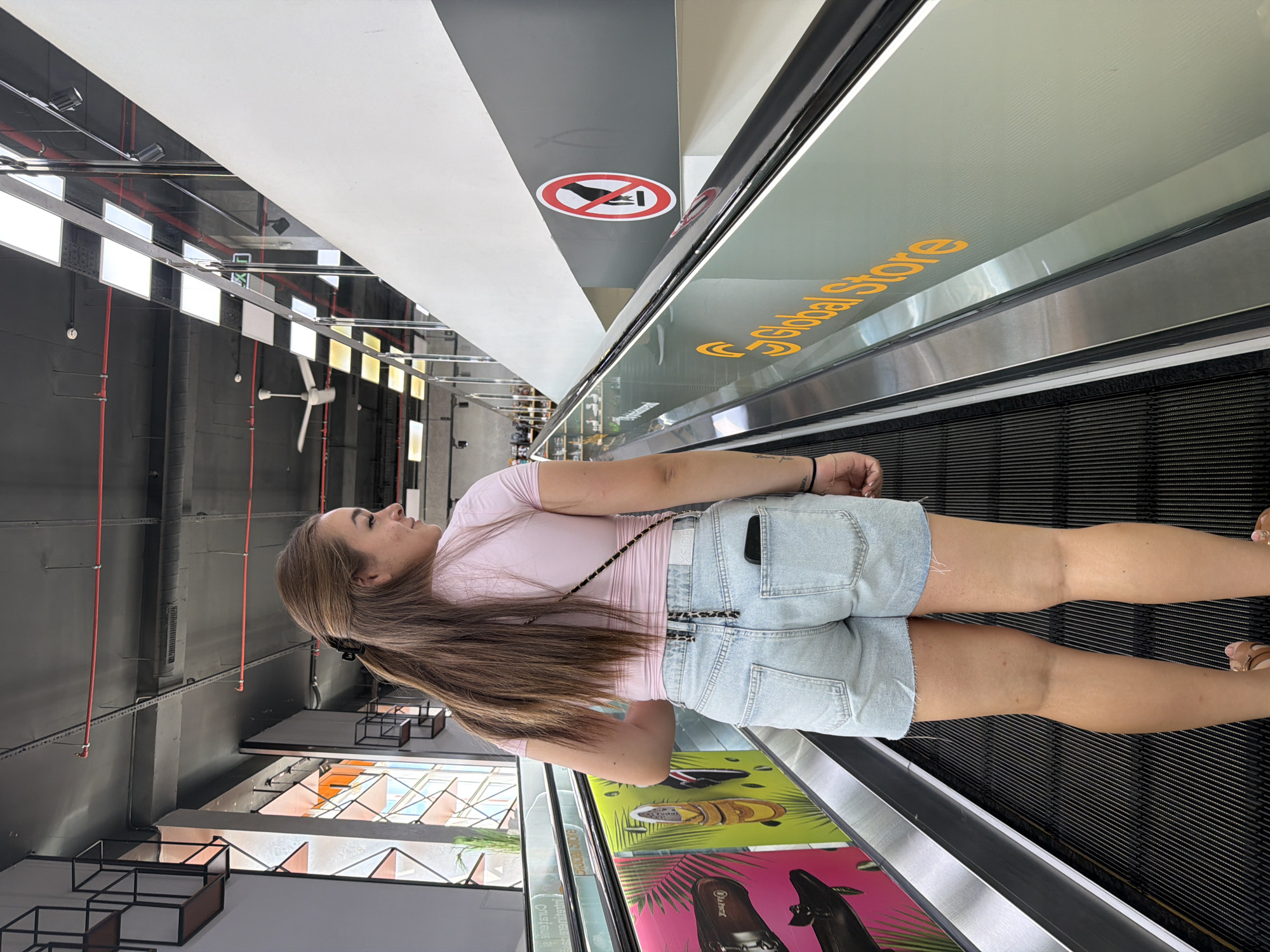 A young woman with long brown hair is ascending an escalator in what appears to be a modern commercial building. She is wearing a pink t-shirt and light-wash denim shorts. Her back is to the camera as she looks over her left shoulder. The escalator has a metallic handrail and a glass barrier. Visible on the barrier are the words "Global Store" in orange lettering and a circular red and white sign depicting a prohibition symbol over a hand. The environment includes a dark ceiling with exposed pipes and lighting fixtures, as well as some shelves and a window in the background. The scene is well-lit, suggesting it is daytime or the interior is brightly illuminated. The woman has a phone in the back pocket of her shorts and a small black bag slung across her body.