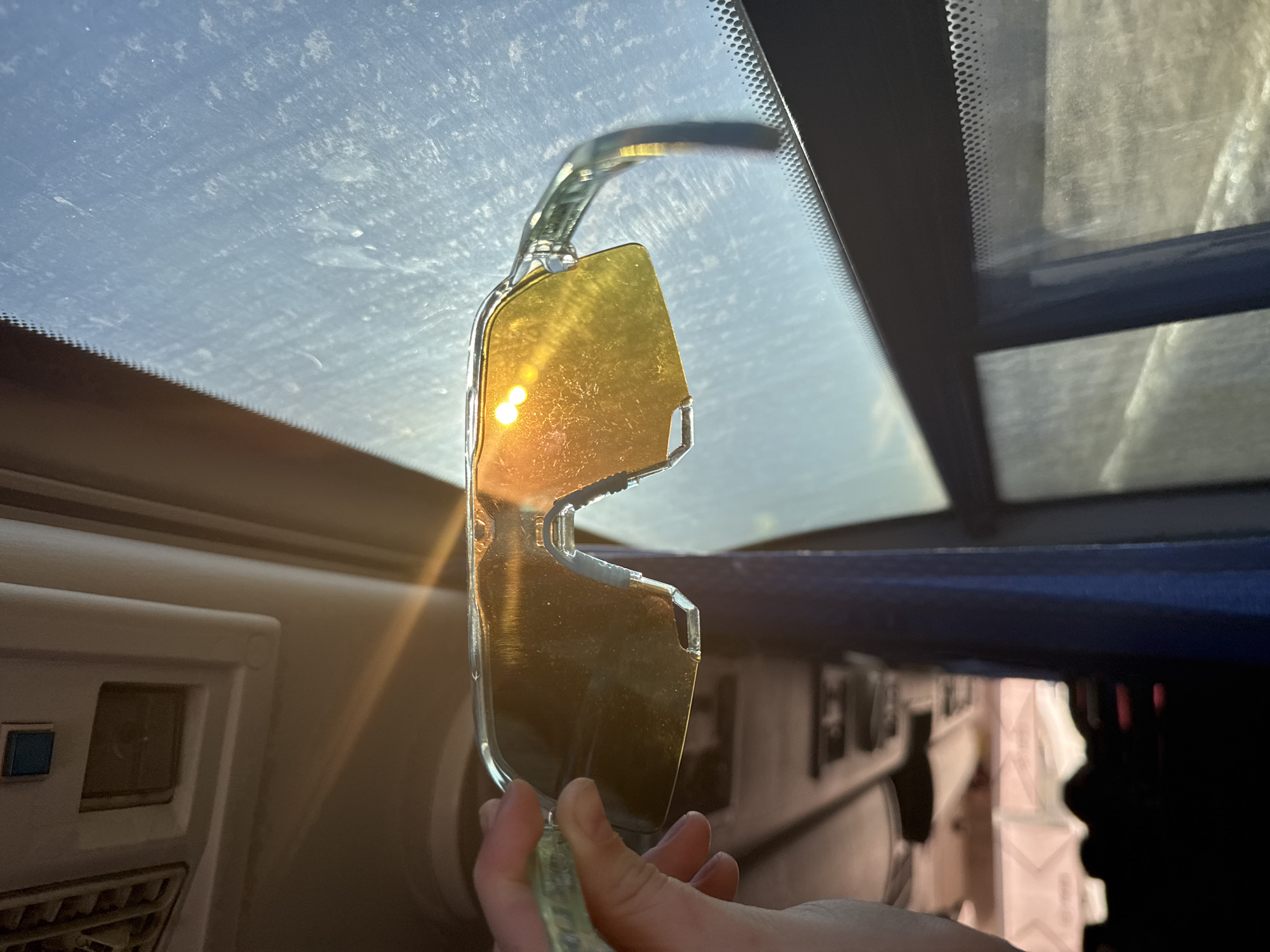 A pair of yellow-tinted sunglasses is held up to a car windshield. The sun is reflecting brightly off the lenses of the glasses. The interior of the car is visible, including the dashboard and the sun visor. The location, Челва, Russia, cannot be determined from this image.