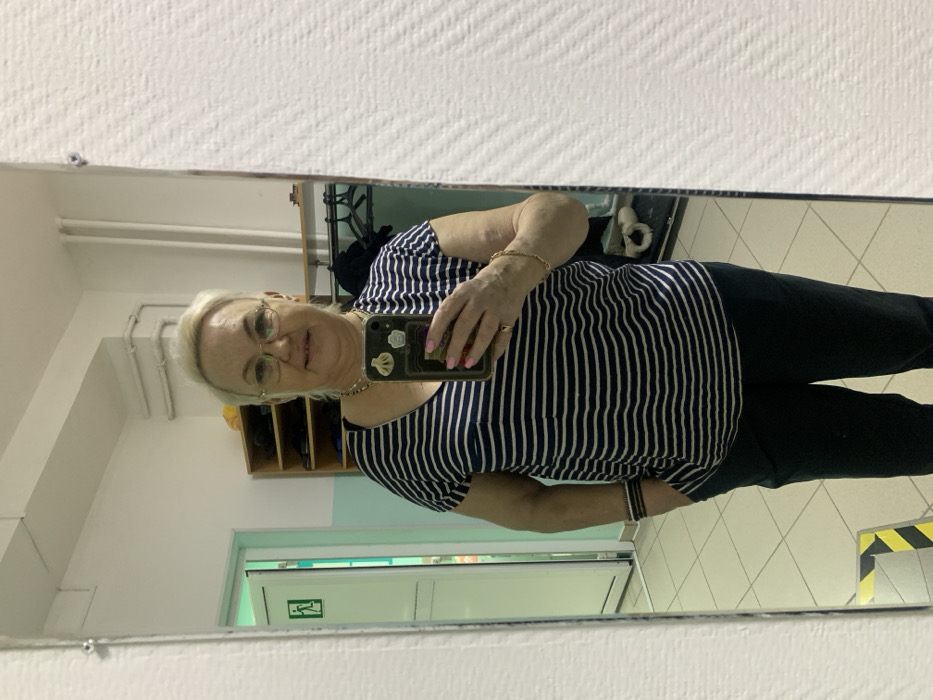 A middle-aged woman is depicted in a full-length mirror selfie taken in an indoor public or commercial setting in Chaikovsky, Russia. The woman, with short blonde hair and spectacles with thin metal frames, is wearing a navy blue and white striped V-neck top and dark trousers. Her right hand holds a smartphone with a light-colored, stickered case, featuring a dumpling motif and a card holder, positioning it to capture her reflection. Her fingernails are painted pink. A gold-colored chain necklace, a gold-colored bracelet on her right wrist, and dark beaded bracelets on her left wrist are visible. A ring is present on her right ring finger. Her left hand is in her trouser pocket. She displays a slight smile. The reflected background reveals light-colored walls and a light-colored tiled floor. To the left, a partially open doorway with a light green frame and a white door displays a green 'running man' emergency exit sign. To the right and behind the subject, a wooden shelving unit and a dark clothing rack with hanging garments are visible. White pipes run horizontally along the upper section of the wall. A yellow and black striped warning tape is present on the floor in the lower-left corner.
