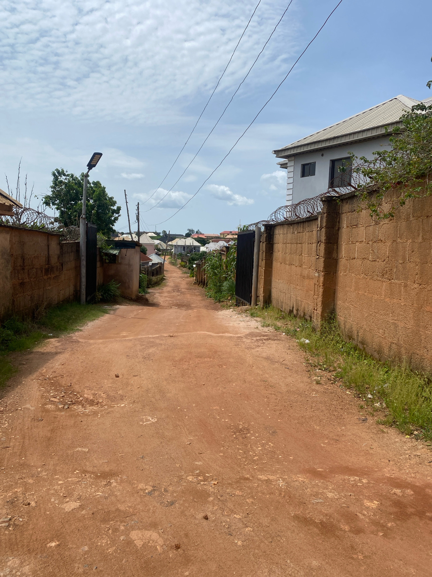 The image depicts a dirt road in a residential area. The road is unpaved, with a reddish-brown surface and scattered stones and debris. On both sides of the road are brick walls topped with barbed wire, suggesting private property. A few trees and bushes grow along the edges of the road. In the distance, houses with various roof styles and some with metal gates are visible. A street lamp stands to the left of the road. Power lines stretch across the sky, which is partly cloudy. The overall impression is of a somewhat rural or underdeveloped urban environment. The scene is likely located in Jos, Nigeria, given the architectural style and the reddish earth. There are no people visible in the image.