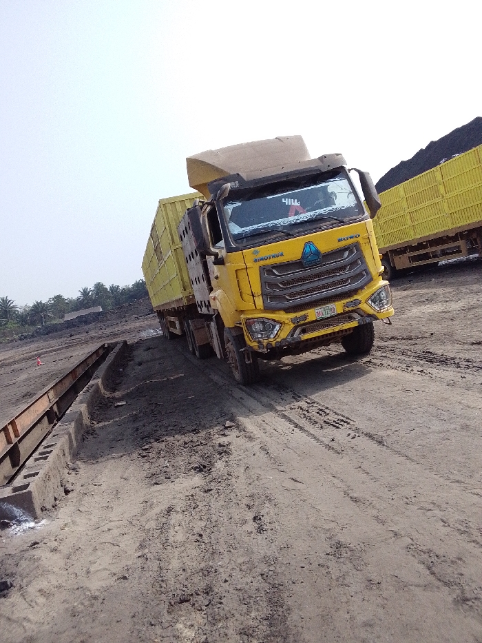 The image depicts an outdoor industrial or mining scene in Hoche, Nigeria, under an overcast or hazy sky. The primary subject is a yellow Sinotruk Howo heavy-duty truck, positioned at an angle on an uneven, dark dirt road with visible tire tracks. The truck features a raised, yellow, high-sided cargo body or trailer, typical for bulk material transport. Its front bumper displays minor deformation, and a Nigerian license plate, "04A12EB," is affixed below the grille.
To the left of the truck, a long, brown, rusty metal structure, resembling a weighbridge or a component of a loading facility, is partially visible, supported by concrete blocks embedded in the dirt. In the background on the right, another yellow truck or trailer, also with a high-sided cargo body, is parked, seemingly laden with a dark, granular material, likely coal or ore, which is also piled in large mounds further behind. Distant palm trees or similar tropical vegetation are visible in the far background on the left horizon. No individuals are discernible in the frame.