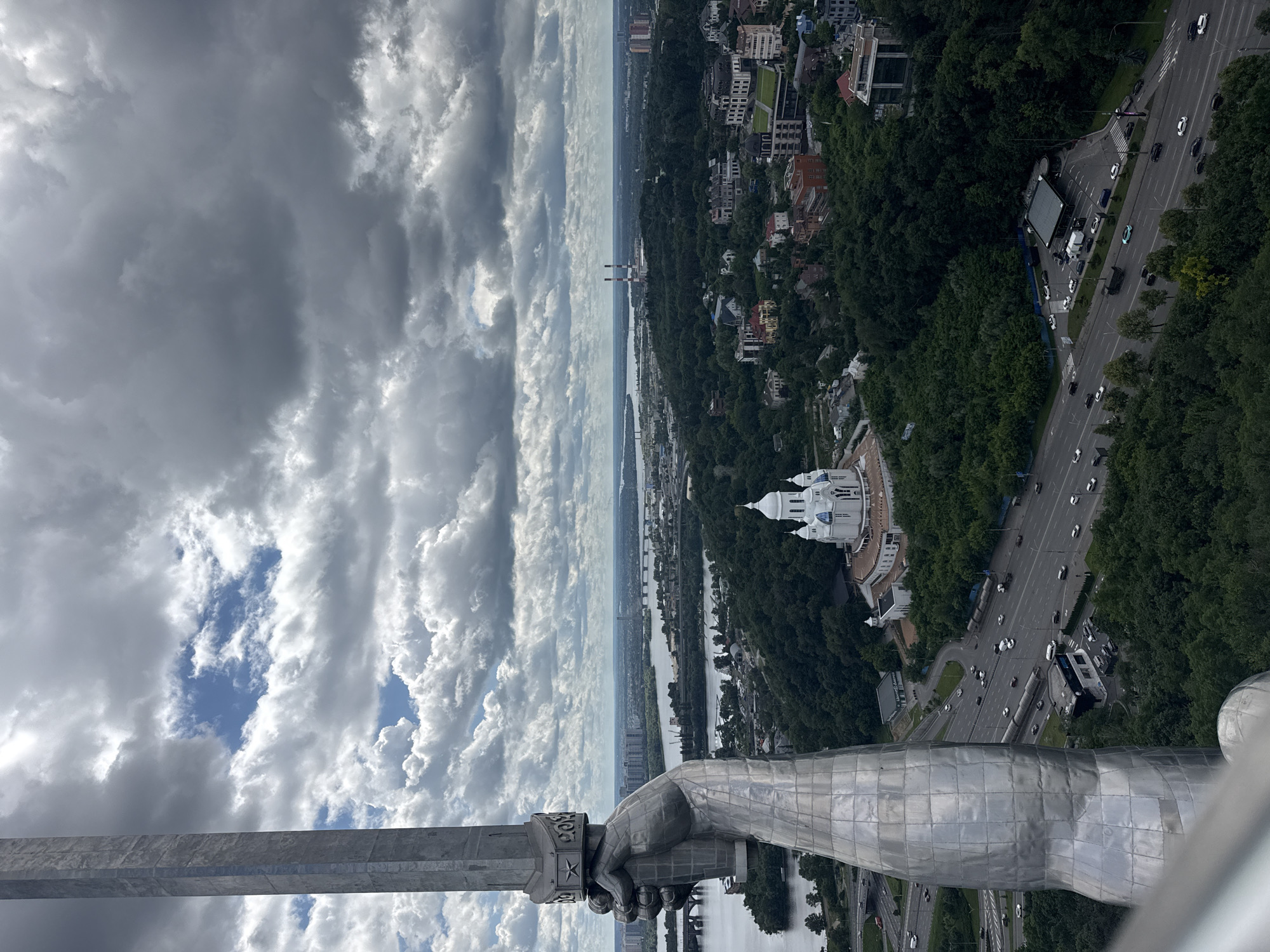 This is a high-angle, exterior, full shot of the Motherland Monument in Kyiv, Ukraine, on a cloudy day. In the foreground, the massive, metallic sculpture of a woman's hand holding a sword dominates the left side of the frame. The sword, a tall, faceted silver object, extends upwards into the sky, its upper portion obscured by the frame. The woman's hand is also made of a reflective, segmented metal, detailed with rivets and seams.
Beyond the monument, a sprawling urban landscape unfolds. A wide river, likely the Dnipro, stretches across the middle ground, with bridges spanning its width in the distance. On the riverbanks and further inland, dense green trees cover rolling hills, interspersed with various buildings. A prominent white church with blue domes stands out amongst the greenery on a hill. Roads with flowing traffic, including cars and trucks, are visible winding through the city and its surrounding forests. In the far distance, industrial structures with smokestacks and urban high-rises complete the cityscape under a dramatic, cloud-filled sky. The overall scene captures a panoramic view of Kyiv from an elevated perspective.