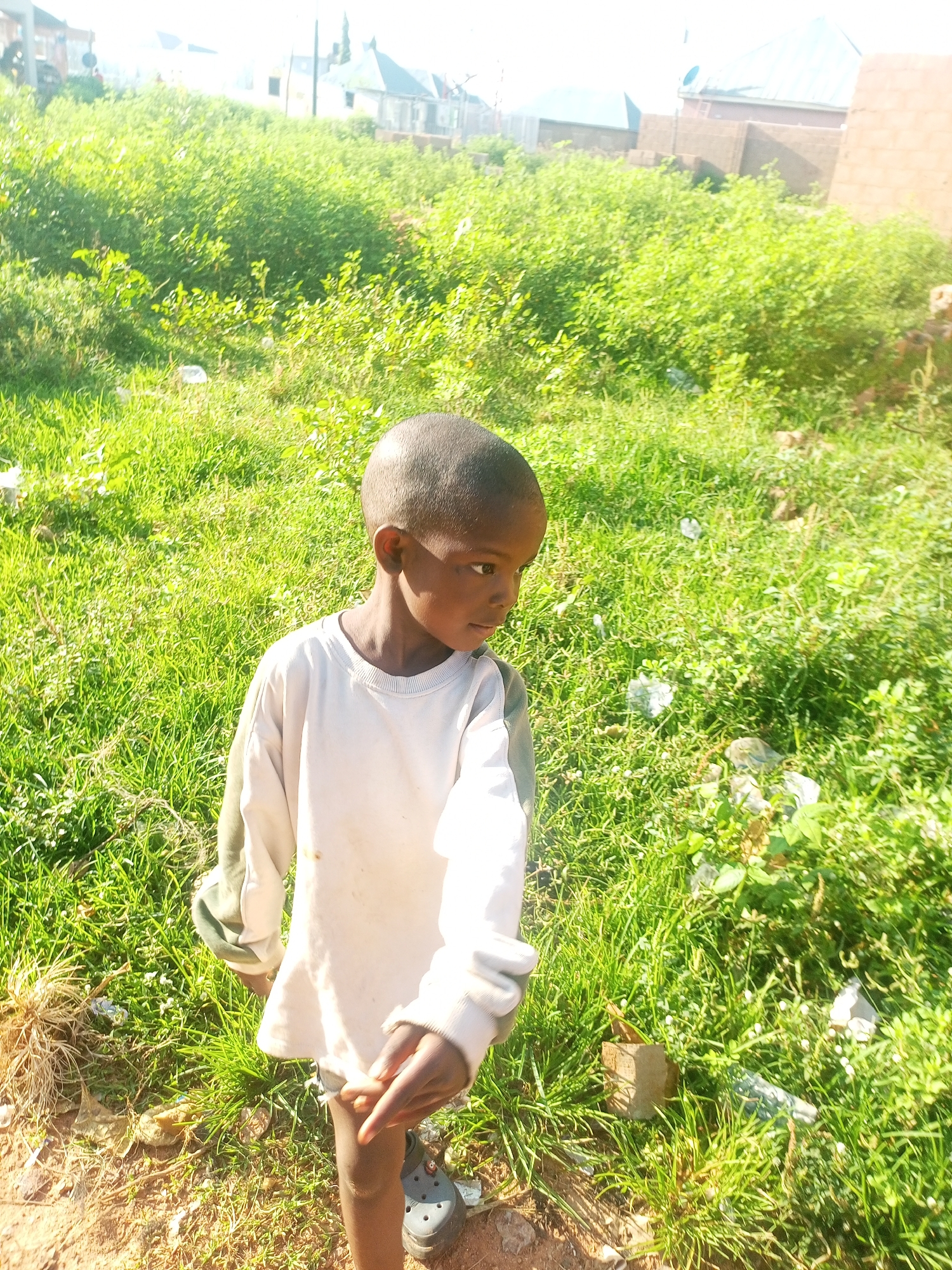 A young boy stands in a sunlit grassy area, possibly a vacant lot or undeveloped land, with buildings visible in the background. The boy is wearing a light-colored long-sleeved shirt and dark shoes. His right hand is slightly raised and open. The ground is covered in green grass and some dry debris. The buildings in the background appear to be residential structures with visible roofs. The lighting suggests it is daytime, with bright sunlight casting shadows. The overall impression is of an outdoor scene in a possibly rural or semi-urban setting within Tempure, Nigeria.