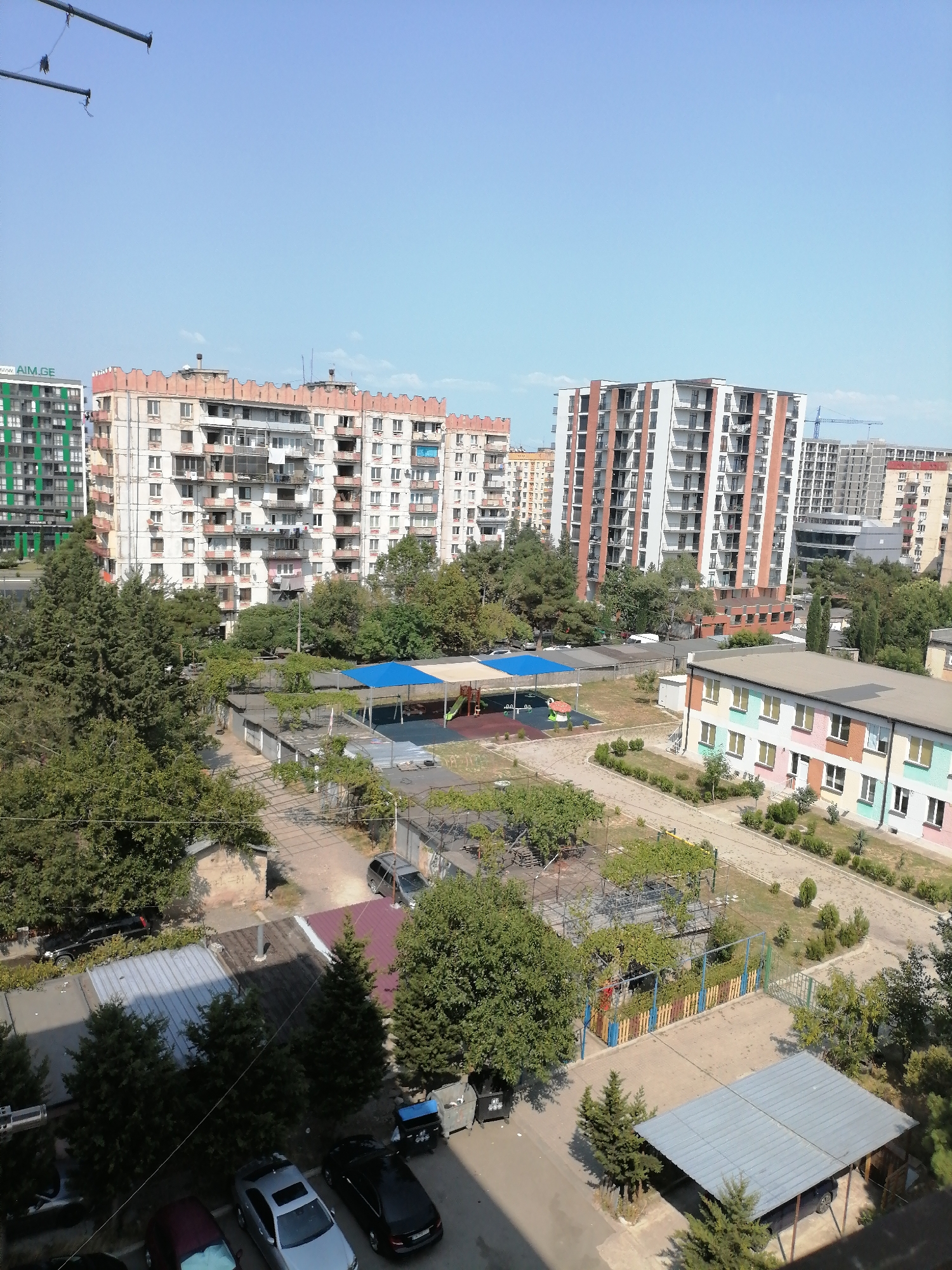 This image captures an urban scene in Rustavi, Georgia, likely from an elevated perspective. The foreground features a parking area with several cars, including a silver sedan and a black sedan parked under a metal carport. Adjacent to the parking area is a small playground with a blue canopy and colorful playground equipment. Beyond the playground, a low-rise building with a colorful facade, painted in pastels like pink, green, and yellow, is visible.
In the midground, a row of garages with flat roofs runs horizontally across the frame. The area around the garages is covered with gravel and sparse vegetation, with some trees and bushes interspersed. Further back, a cluster of trees creates a green canopy.
The background is dominated by multi-story apartment buildings and some construction sites. On the left, a tall, modern green apartment building with numerous balconies stands out. To its right, older, pink-toned apartment buildings with a distinctive crenellated roofline are visible, followed by a very tall, modern residential building with white walls and brick accents. In the distance, a construction crane indicates ongoing development. The sky is clear and blue, suggesting a sunny day. The overall impression is of a residential neighborhood with a mix of older and newer architecture, green spaces, and active urban life.