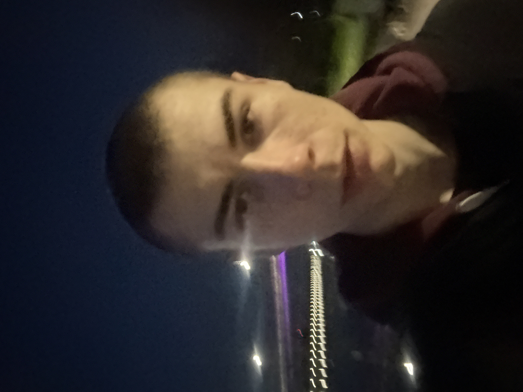 A blurry selfie taken at night. A person with short hair and light skin is in the foreground. The background features blurred lights and a dark sky. The lights suggest a cityscape. Based on the faint purple and white vertical lights, it is likely taken facing the city of Plovdiv, Bulgaria. The subject is likely near a bridge or elevated area overlooking the city. The photo is taken at night, as indicated by the dark sky and artificial lighting.