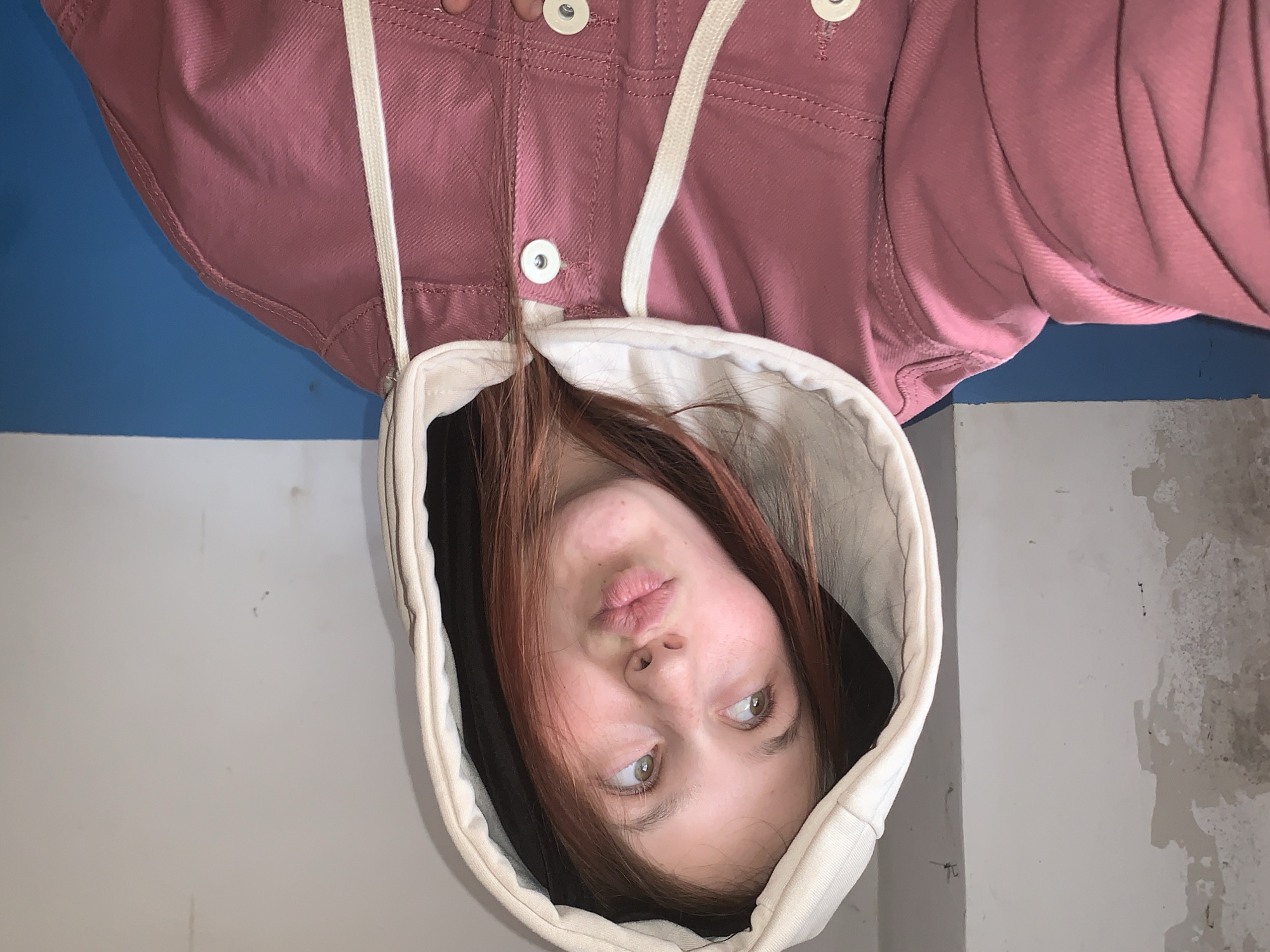 An inverted portrait features a young person, likely female, with reddish-brown hair, positioned upside down. She looks upwards with a pensive or slightly pouting expression, her lips gently pursed. She is wearing a dusty rose or mauve-colored jacket with white buttons and white drawstrings. An off-white or cream-colored hood, possibly part of a separate garment worn underneath or a two-tone jacket, frames her face.
The background is an indoor wall, horizontally divided into two sections. The upper section, behind the person's torso, is a solid blue. The lower section, behind her head, is a light grey or off-white, displaying noticeable wear and texture, with areas of distressed or peeling paint visible, particularly on the right side. The lighting appears even. There is no visible text in the image. The scene is set in Фоки, Russia.